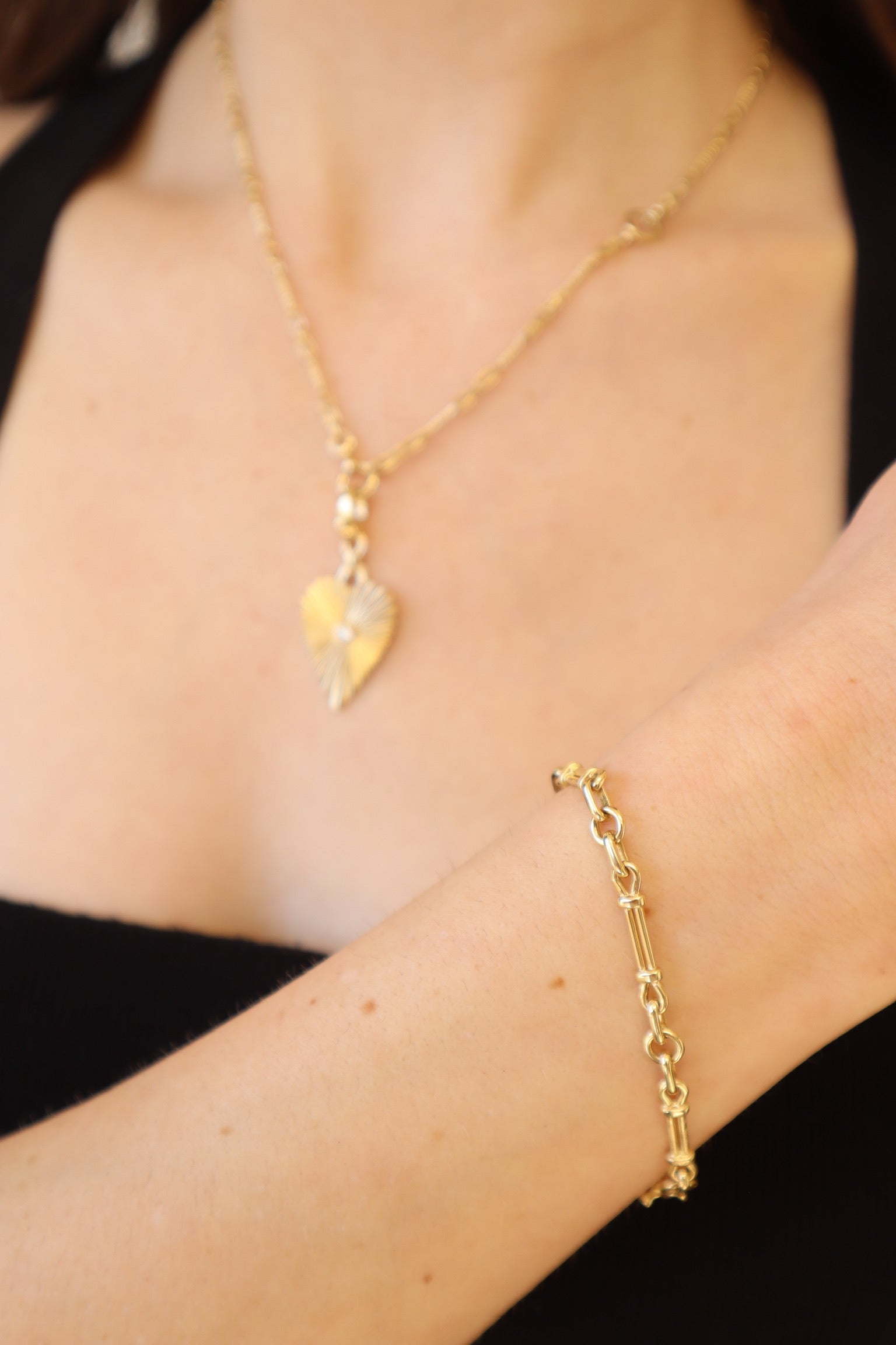 The Lisbon Bracelet by Janet Heller Fine Jewelry is a gold chain-link design with small loops and elongated cylindrical segments. It features a clasp adorned with a small diamond accent. The bracelet can be worn on its own or stacked with other bracelets.