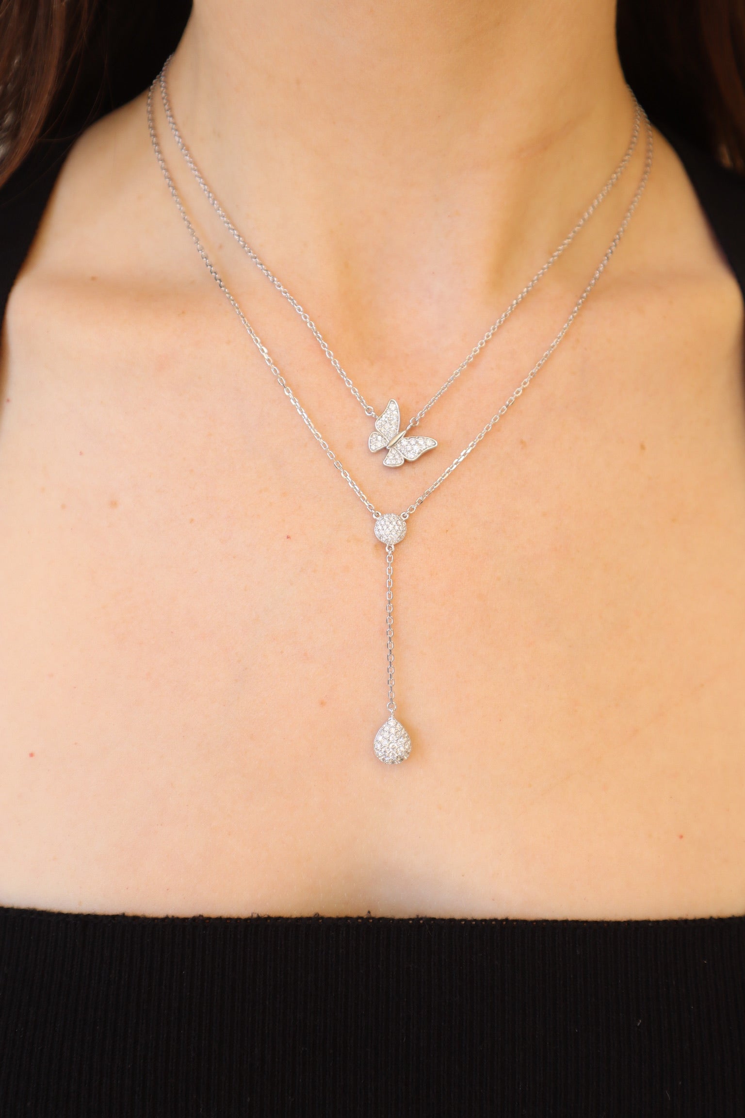 A person in a black top flaunts the Mini Sideways Butterfly Necklace with layered silver chains, featuring a butterfly pendant and a lariat ending in heart and round charms encrusted with small crystals. Customizable options offer a personalized touch.