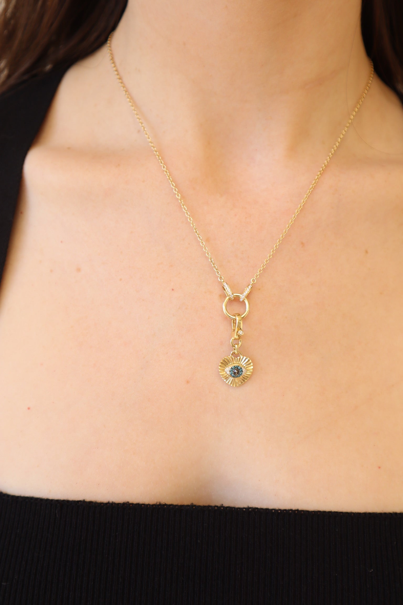 A close-up shows someone wearing the Ribbed Diamond Heart Evil Eye Charm by Janet Heller Fine Jewelry—a necklace with a blue-centered circular pendant. The black top enhances its elegance, set against a softly blurred background that highlights the charms appeal.
