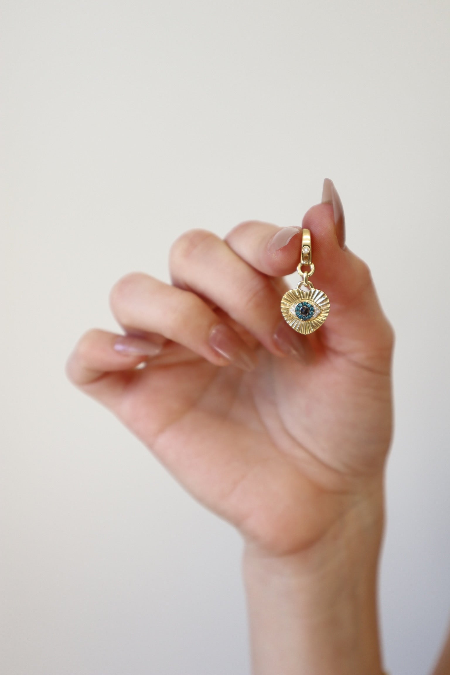 A hand with manicured nails holds the Ribbed Diamond Heart Evil Eye Charm from Janet Heller Fine Jewelry, showcasing an intricate sunburst design and central deep blue gemstone. The piece is set against a plain white background, embodying the timeless allure of expertly crafted treasures.