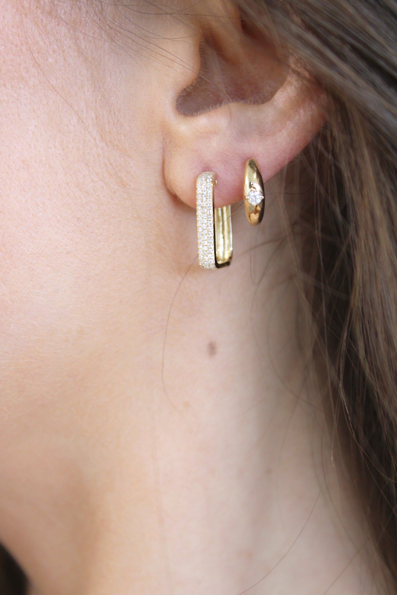 A close-up of a womans ear displays the Janet Heller Fine Jewelry Solitaire Diamond Dome Huggies. Her hair gently drapes, partially covering the outer ear as she wears these exquisite gold hoops encrusted with diamonds.