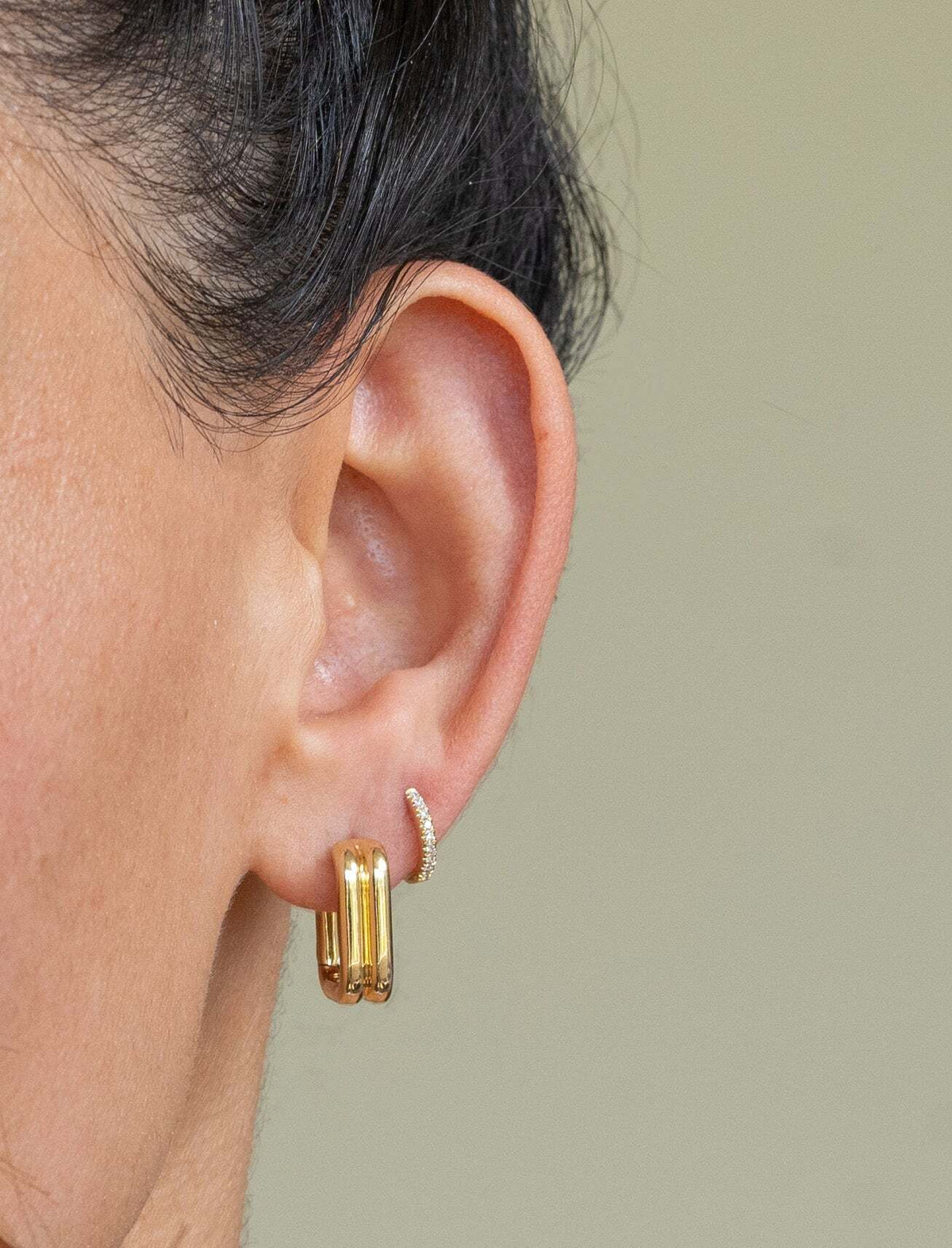 Close-up of a person’s ear wearing Janet Heller fine jewelry: the Mini Diamond Anika Huggies, a curved, diamond-studded stud in the upper lobe and a thick gold hoop in the lower lobe, set against a plain beige background. Perfect for custom jewelry inspiration.