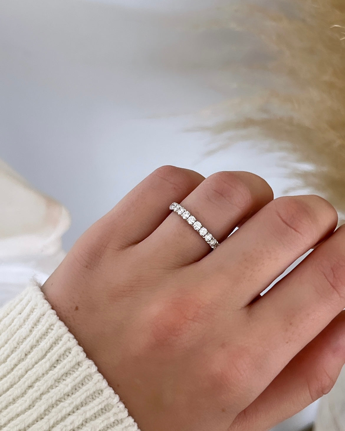 A hand in a white knitted sleeve, graced by the 1ct Round Diamond Eternity Ring on the ring finger, rests gently against a soft, blurred beige background, highlighting the elegance of 18KT gold.