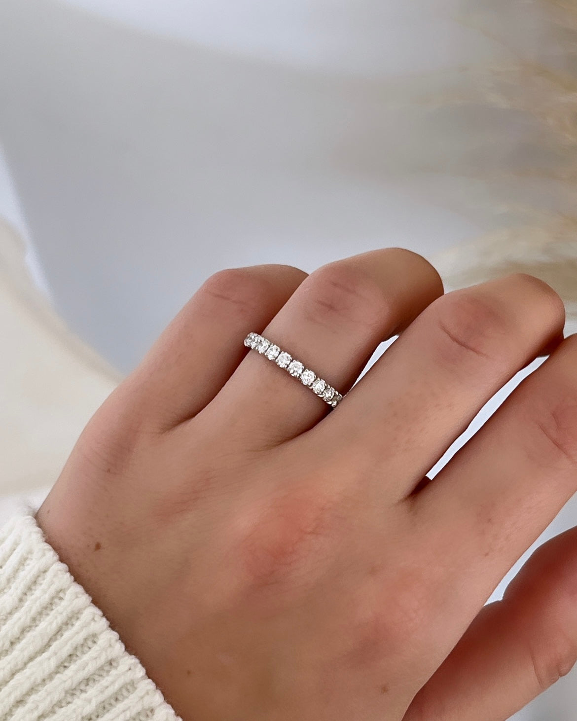 A hand wearing the 1ct Round Diamond Eternity Ring made of thin 18KT gold with VS clarity diamonds rests on the ring finger. The person is in a white, ribbed sweater against a softly blurred, neutral-toned background.