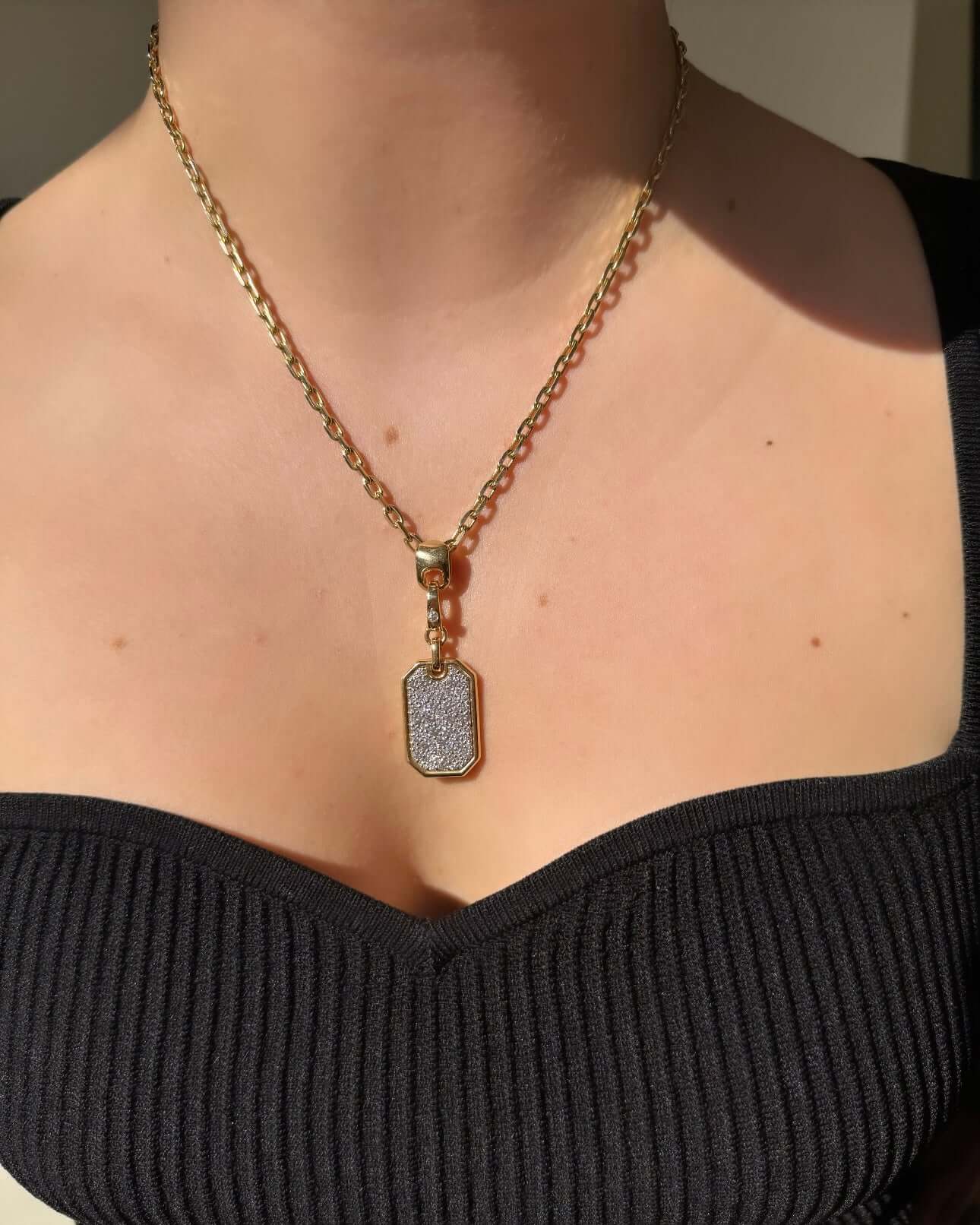 Wearing a gold chain necklace with a Medium Diamond Dog Tag Charm, featuring a textured gray surface, they pair it with a black, ribbed off-the-shoulder top. The well-lit setting accentuates the charm from Janet Heller Fine Jewelry, renowned for its elegant customizable pieces.