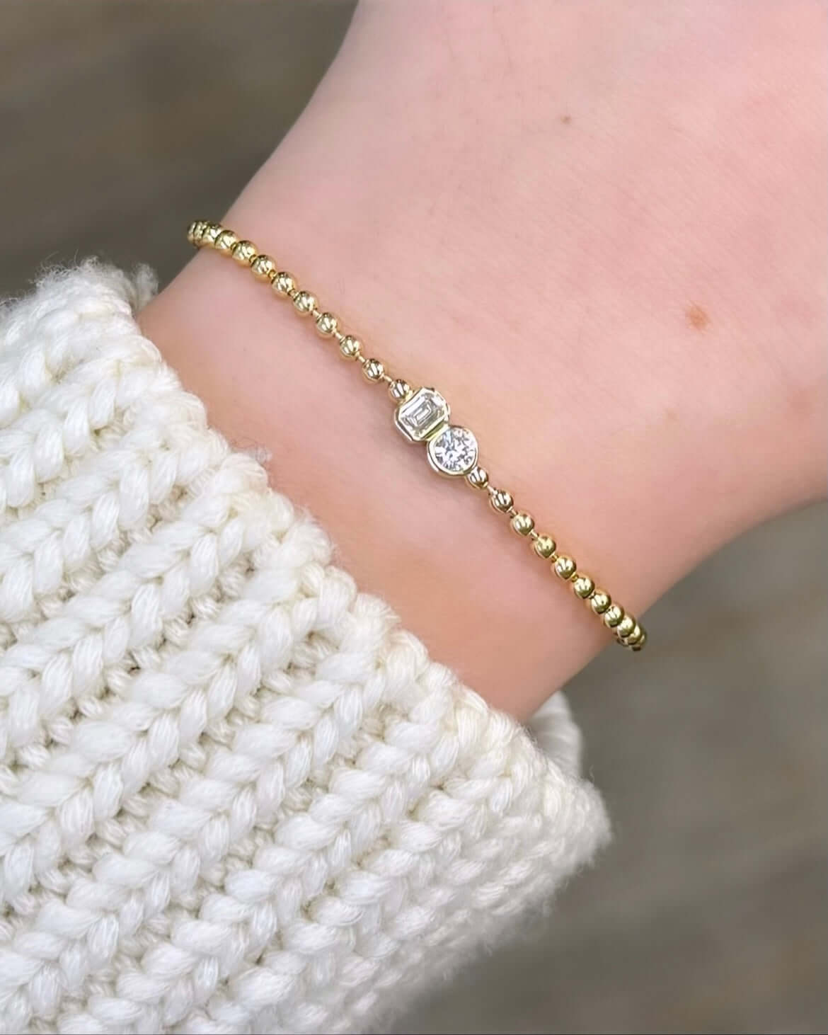 A hand wears the Emerald & Round Toi Et Moi Bracelet with gold beads and assorted gemstones, elegantly gracing a wrist over a cream-colored knitted sweater sleeve, weaving its own love story.