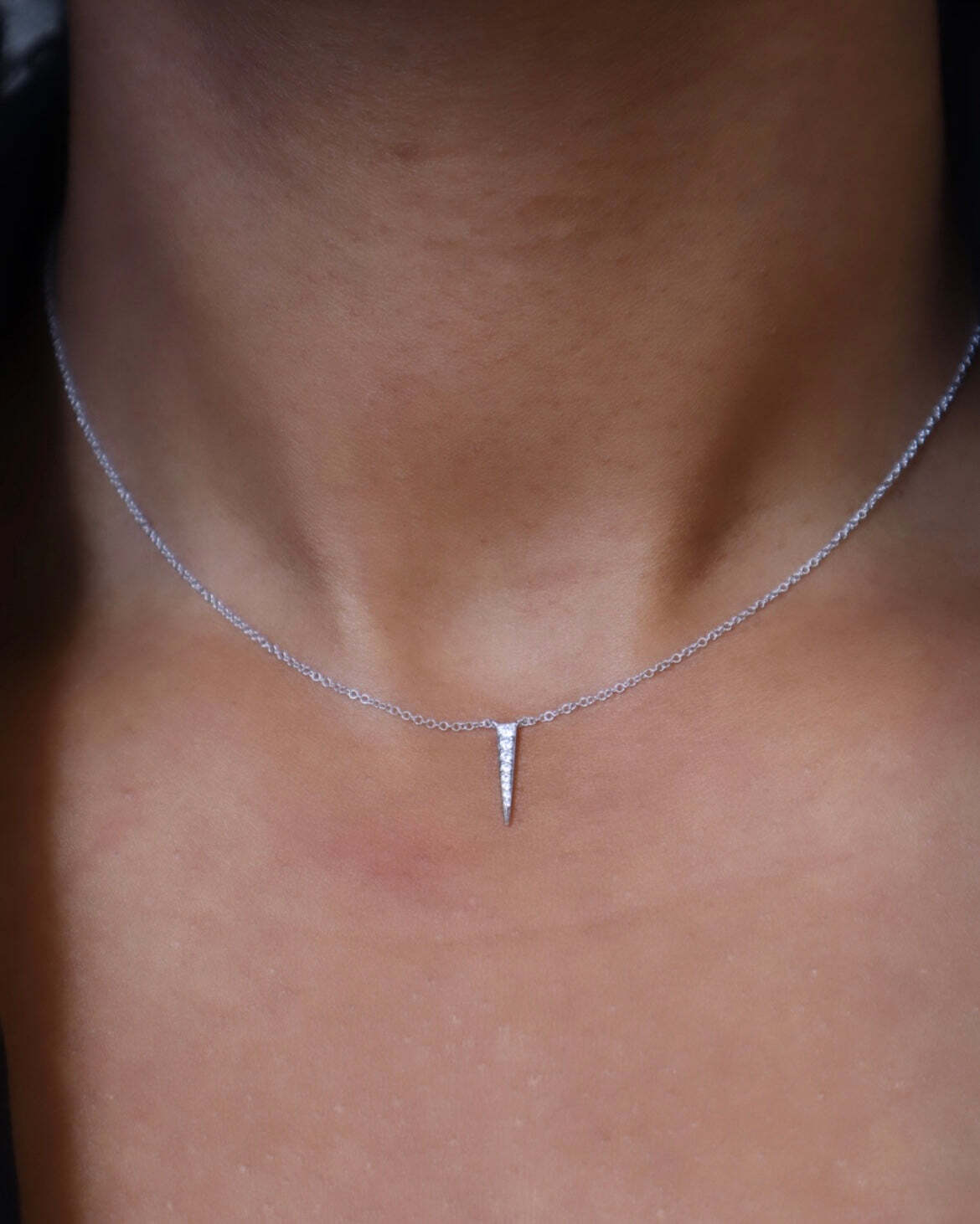 A close-up of a person’s neck wearing the 14KW Mini Diamond Dagger Necklace from Janet Heller Fine Jewelry, featuring a delicate silver chain and a small, pointed dagger pendant at the center.