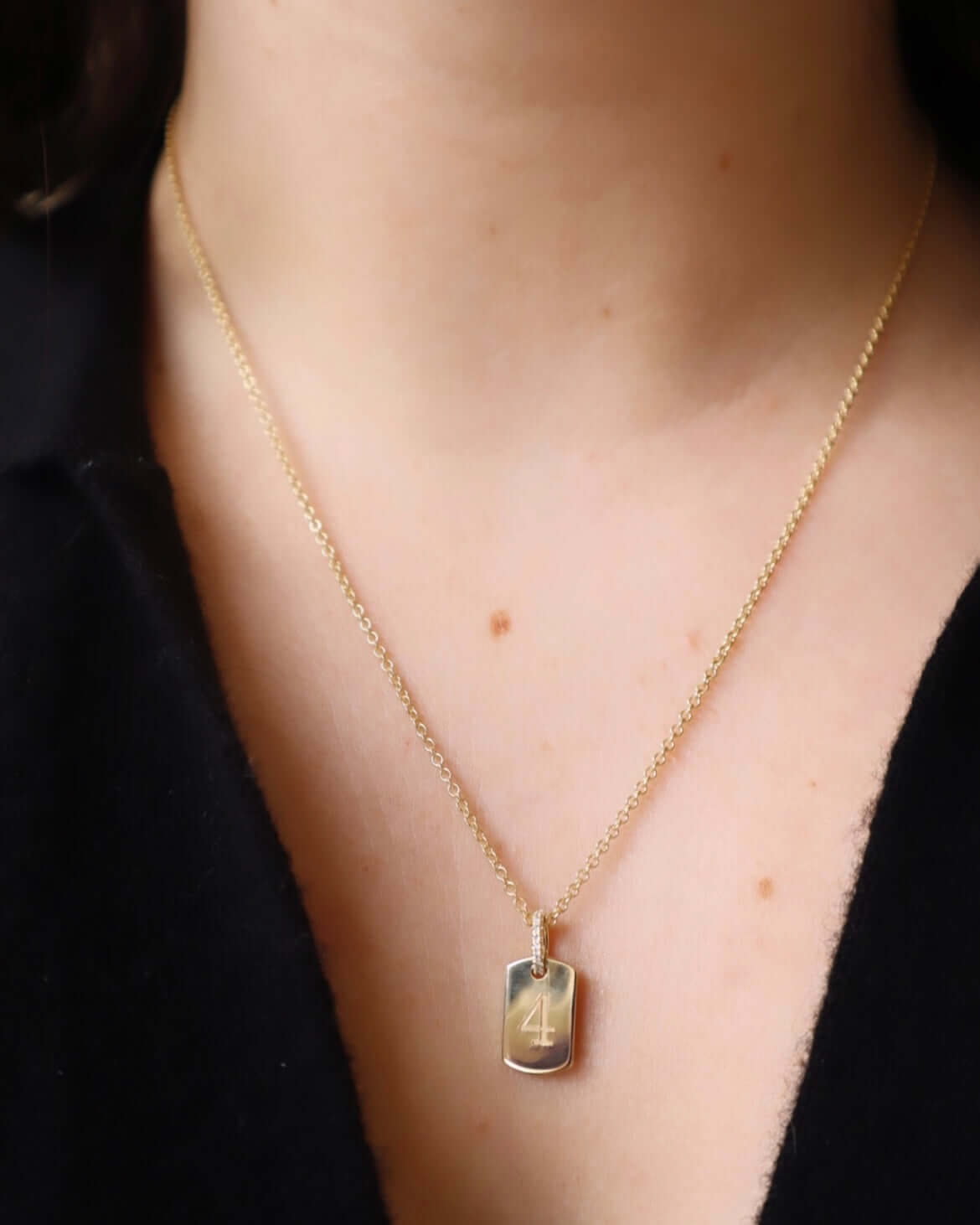 A person in a black shirt wears a Mini Dog Tag Engravable Pendant Necklace from the Sketch Collection, featuring a rectangular pendant engraved with the number 4, creating a personalized keepsake.
