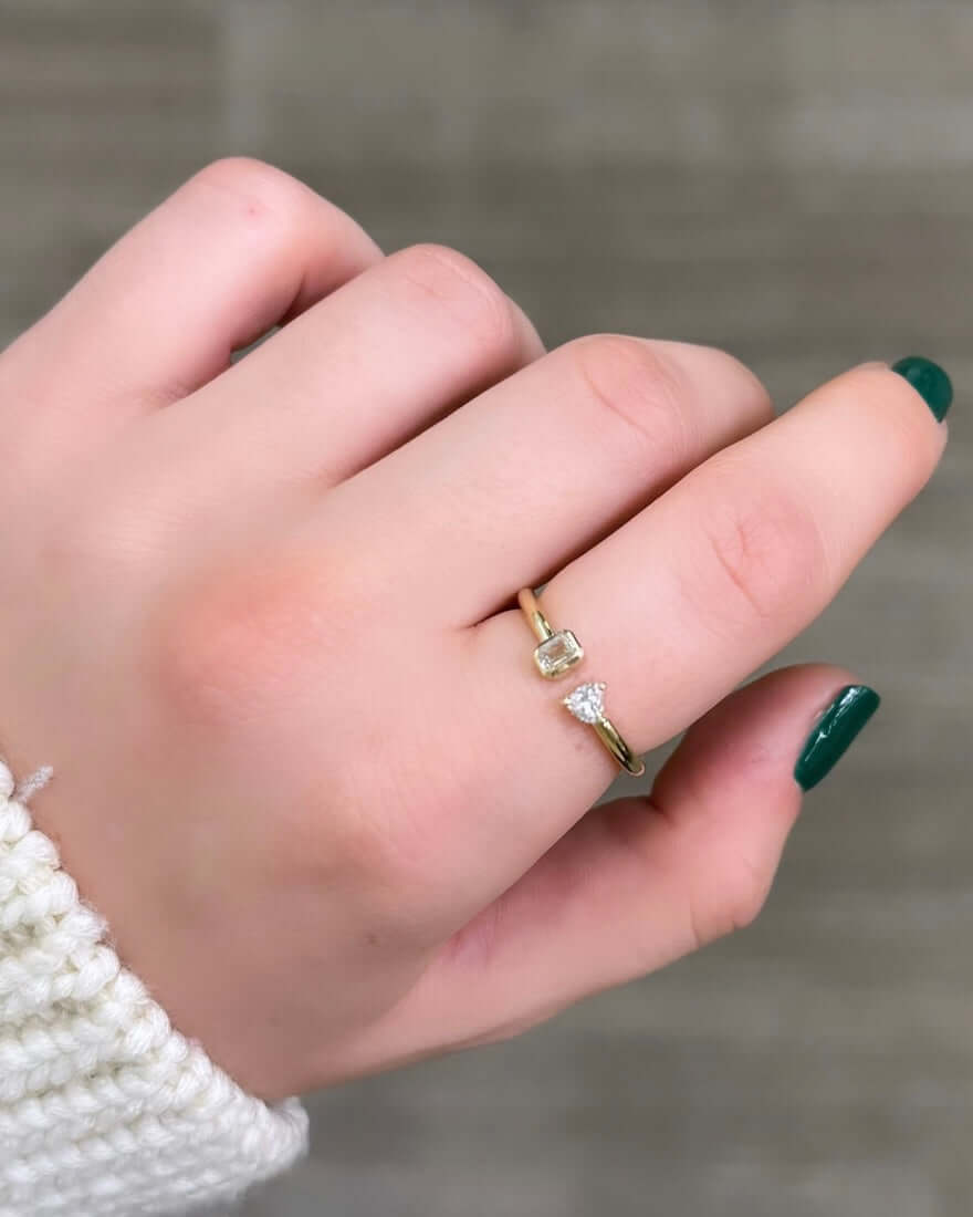 A close-up of a hand with green nails showcases the Emerald & Heart Toi Et Moi Ring, featuring two small gemstones, one clear and one light yellow. The hand elegantly rests against a neutral background, symbolizing timeless love stories.
