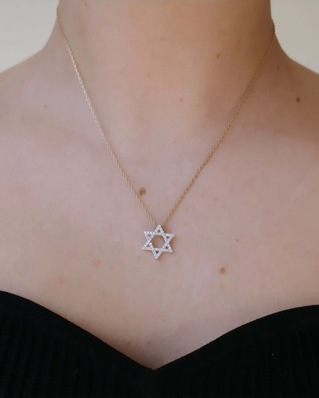 A person adorned with the Diamond Small Star of David Necklace set against a soft-focus, neutral background, elegantly dressed in a black, off-shoulder top.