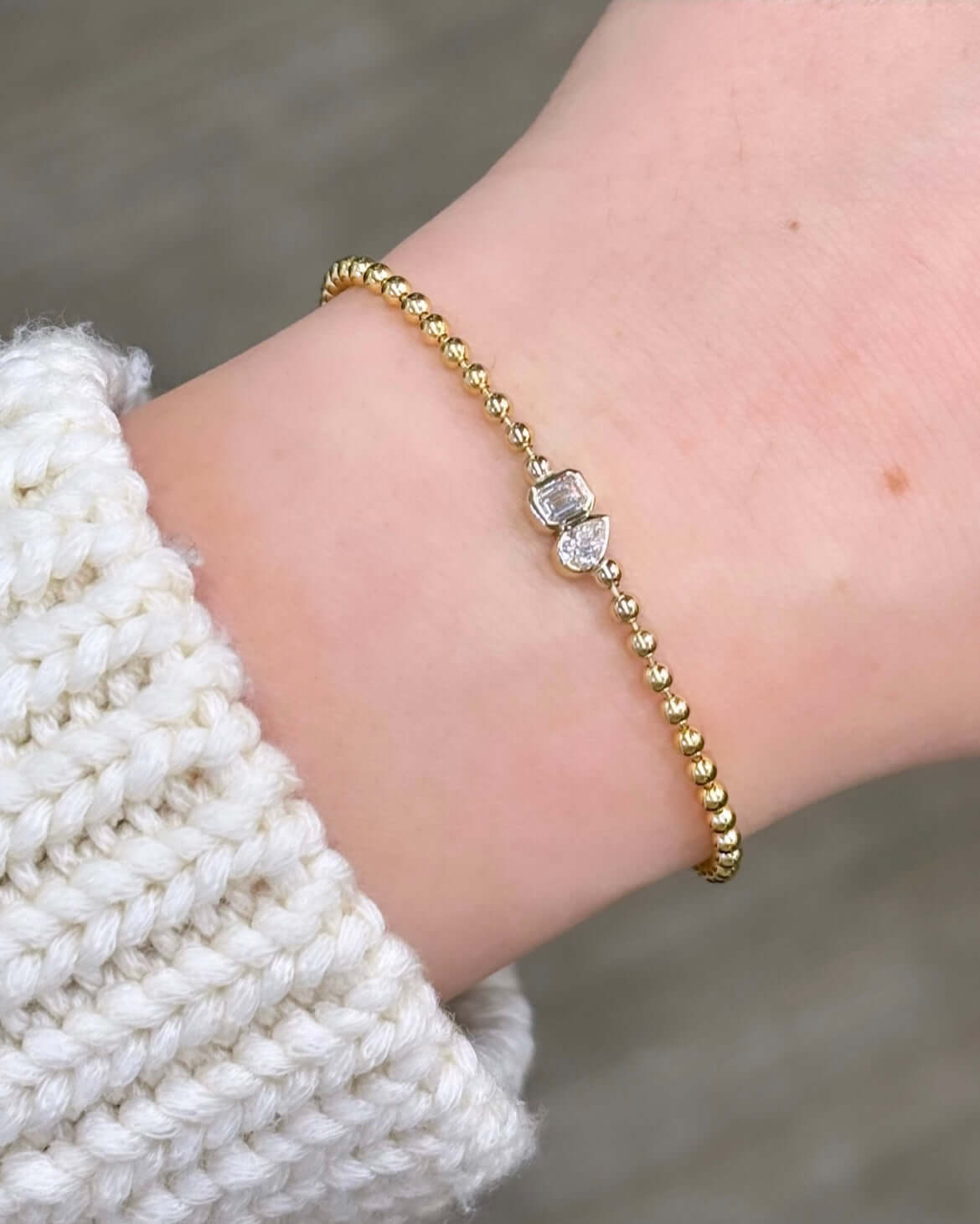 A gold beaded bracelet, the Emerald & Pear Toi Et Moi, with a clear gemstone at its center, symbolizes love against a background of a cream-knit sweater.