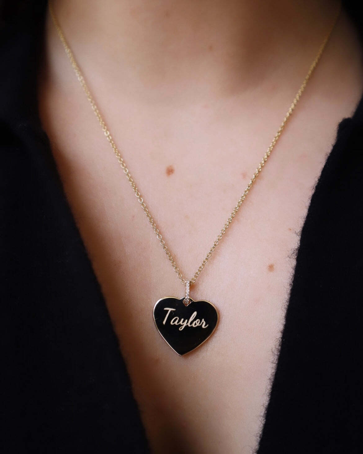 A close-up of a person wearing the Large Heart Engravable Pendant Necklace—a gold chain featuring a black, heart-shaped pendant engraved with the name Taylor—showcases personalized jewelry at its finest. The individual is dressed in a dark-colored, open-collar top.
