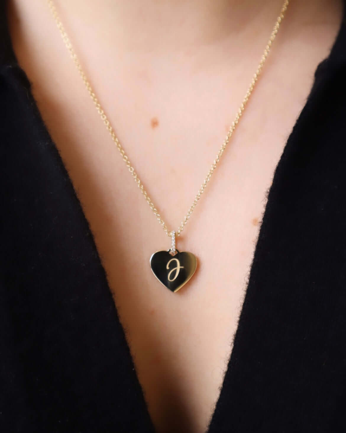A person adorns a Mini Heart Engravable Pendant Necklace, boasting a gold chain with a black heart-shaped pendant embossed with a gold J. It elegantly complements the black garment, evoking the Sketch Collections charm.