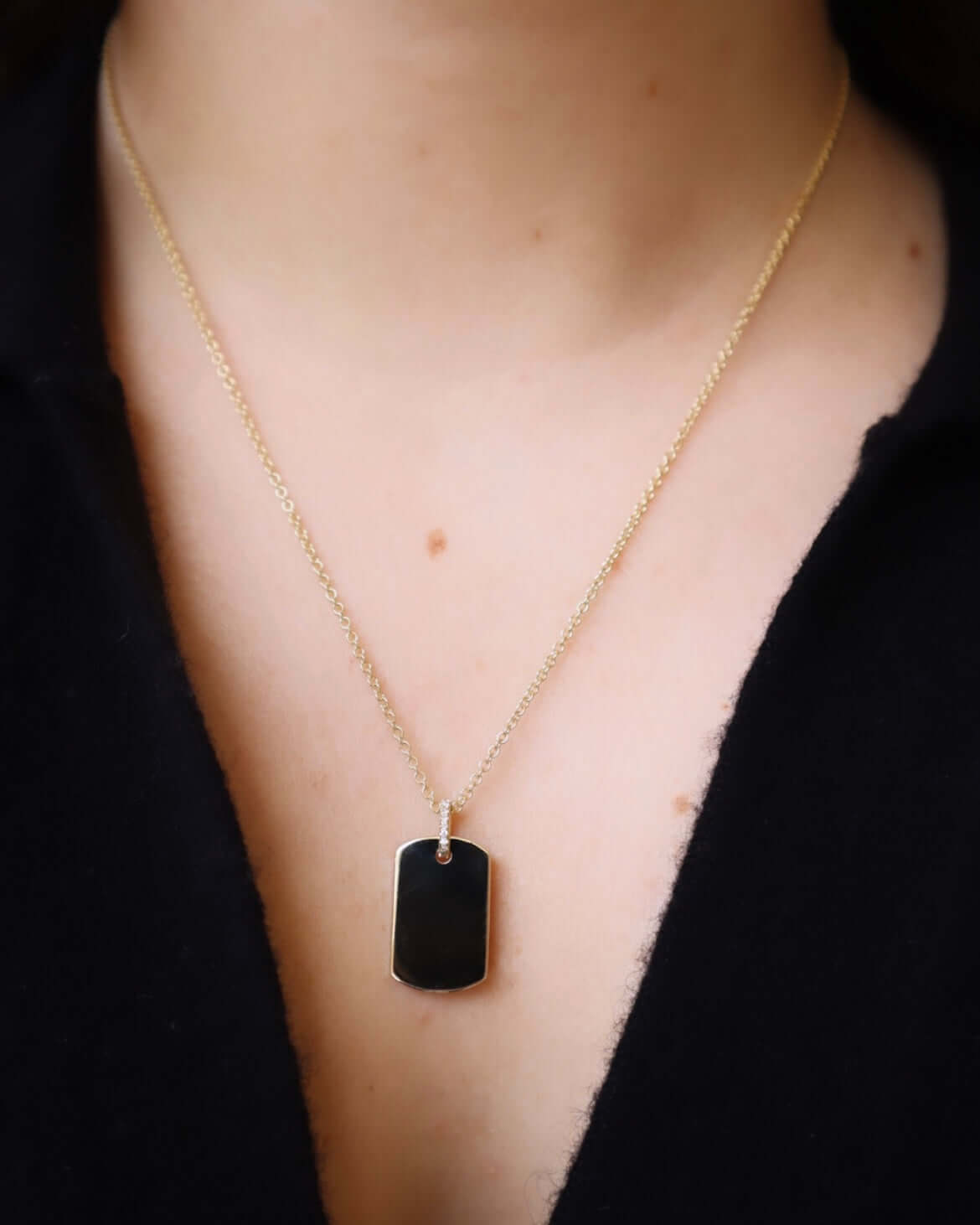 Someone is wearing the Large Dog Tag Engraveable Pendant Necklace, a gold chain with a rectangular black pendant featuring an engraved message, hanging at the center of their chest over a dark shirt. The background is blurred, emphasizing this personal touch.