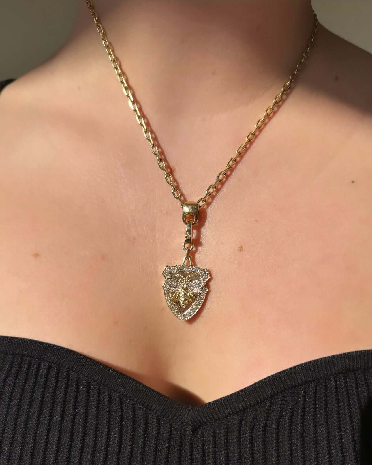 A person elegantly accessorizes a black off-the-shoulder top with a fine gold chain necklace from Janet Heller Fine Jewelry. The centerpiece is the intricately designed Diamond Queen Bee Charm, shaped like a shield and featuring exquisite customizations, complemented by unique jewelry charms.