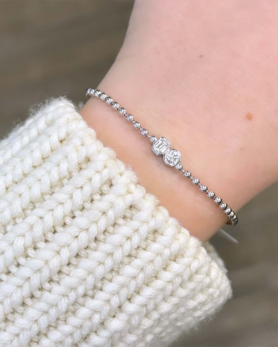 The wearer sports a chunky, cream-colored knit sweater against a blurred, neutral-toned background, their wrist adorned with an Emerald & Round Toi Et Moi Bracelet featuring a central round diamond reminiscent of timeless love stories.