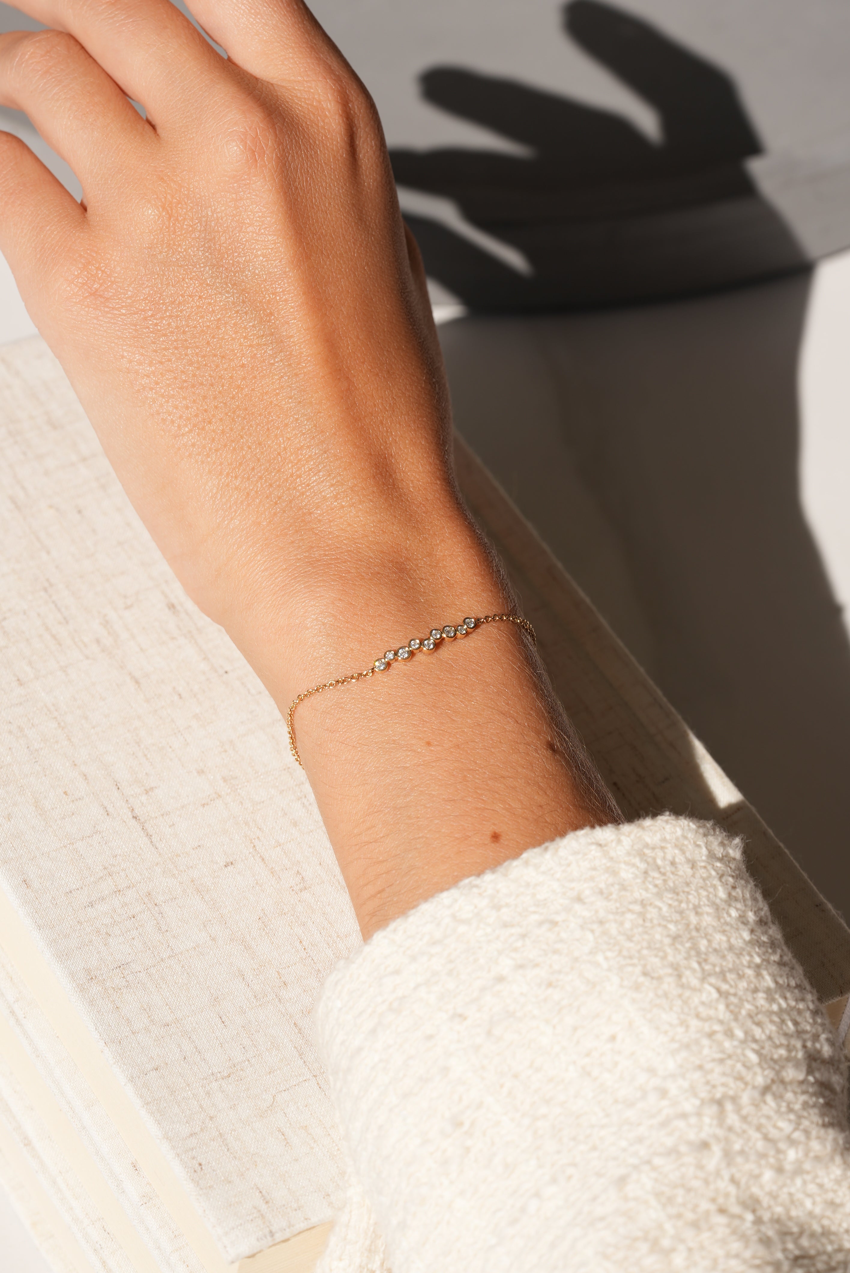 A hand in a cream textured sleeve rests on a white book, adorned with the 9 Diamond Layla Bracelet from Janet Heller Fine Jewelry. Its small sparkling stones glimmer as sunlight casts soft shadows across the elegant scene.