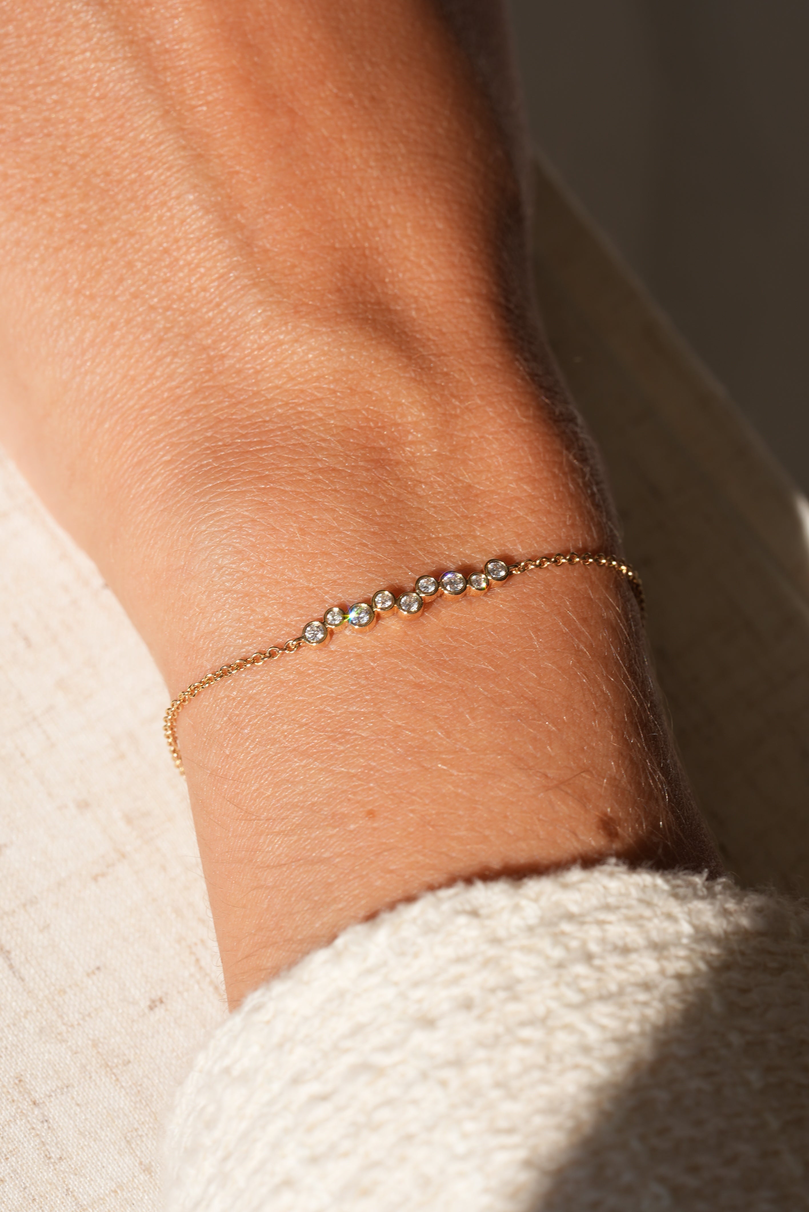 A 9 Diamond Layla Bracelet by Janet Heller Fine Jewelry, crafted in 14KT gold and set with nine sparkling diamonds, adorns a wrist in natural sunlight beside a cream textured sleeve.