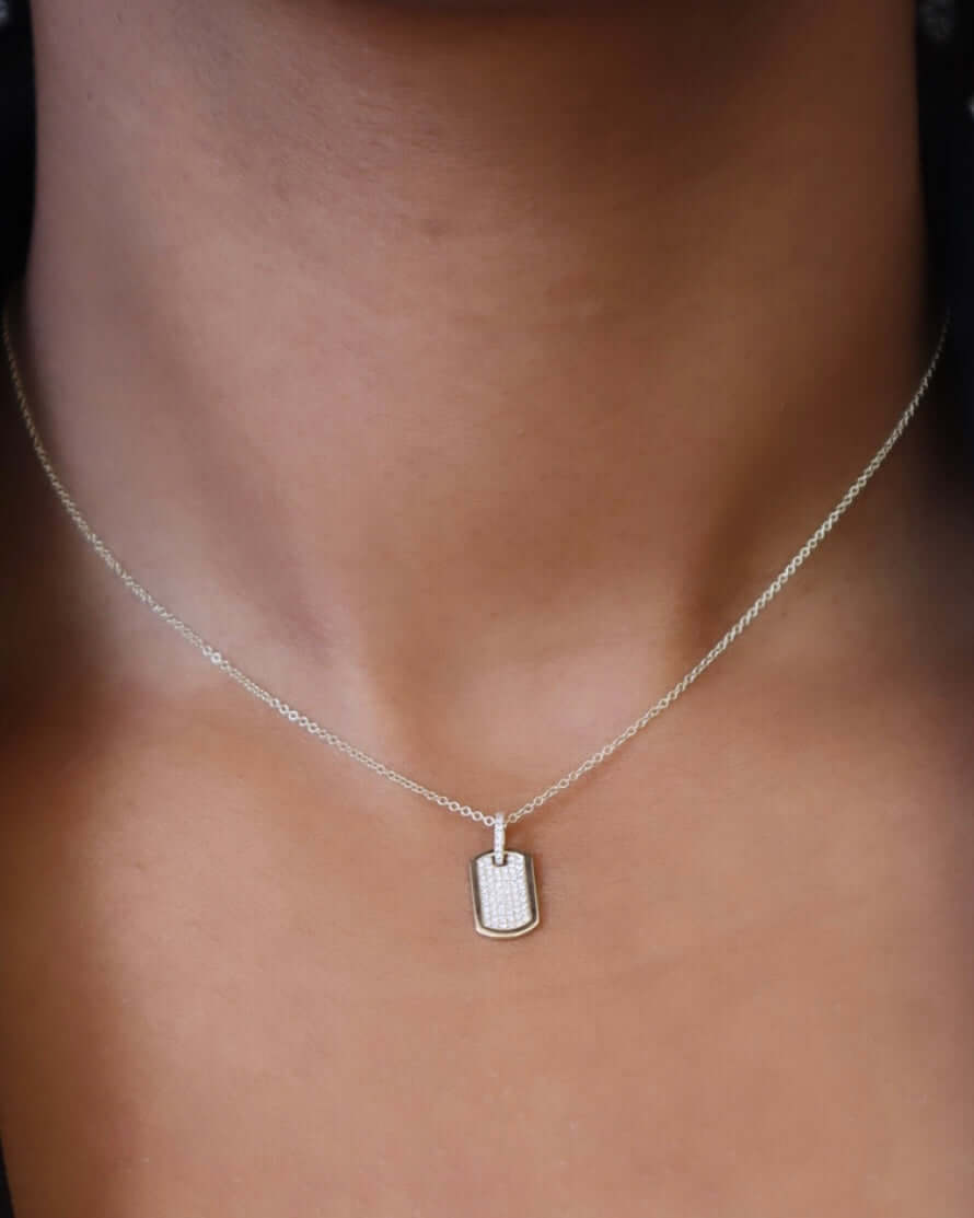 Close-up of a persons neck with a Janet Heller Fine Jewelry Mini Diamond Dog Tag Necklace in silver, featuring a small rectangular pendant. The textured pendant is elegantly centered on the skin against an out-of-focus background.