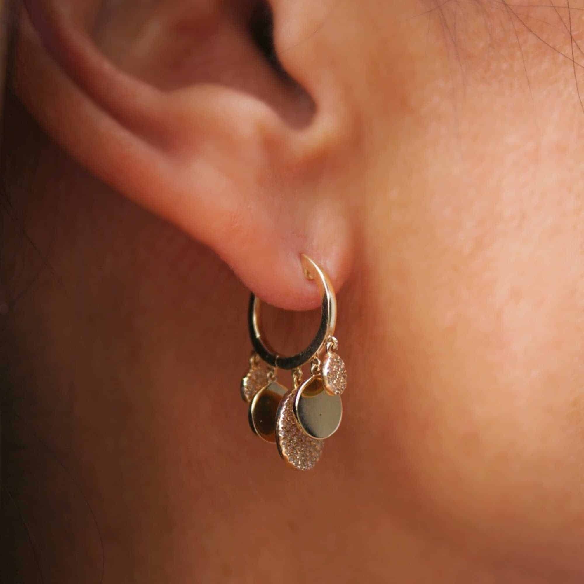 A close-up of an ear wearing the Alternating Diamond Disc Huggies, featuring a gold hoop adorned with multiple small, textured and smooth diamond-accented gold charms that beautifully showcase the artistry of Janet Heller fine jewelry. For custom jewelry or inquiries about this piece, please contact us for more information.