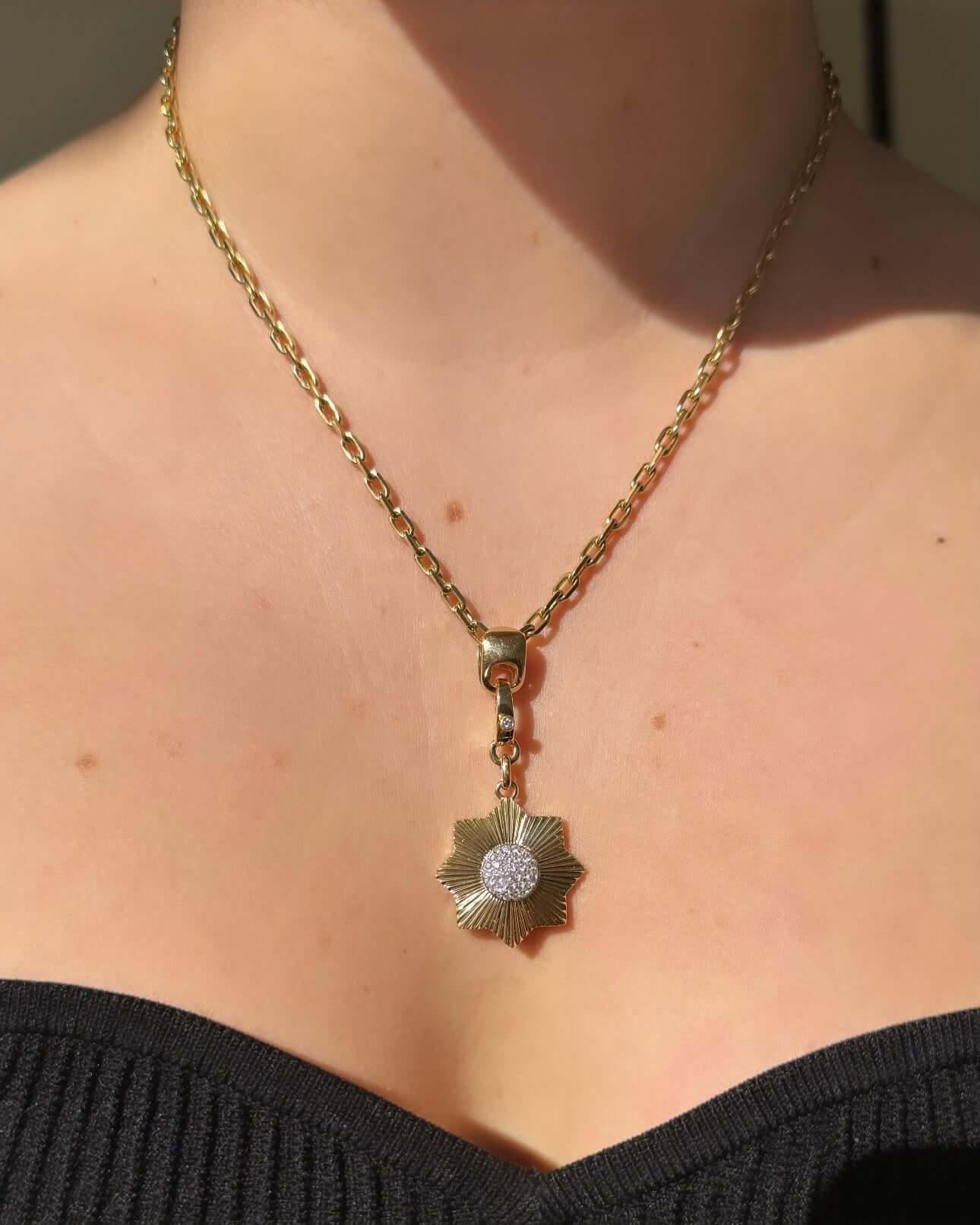 A person adorned with a Pave Diamond Sun Flower Charm necklace glistens under bright lighting, wearing a black, off-the-shoulder top that exudes charm and elegance.