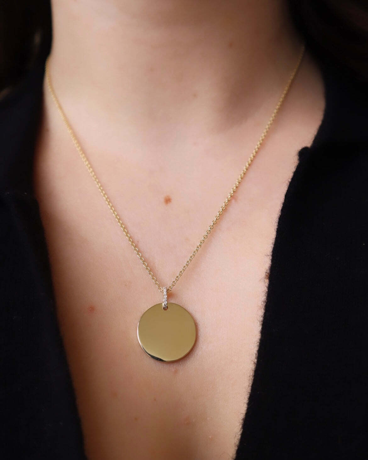 A close-up of a person wearing the Large Round Engravable Pendant Necklace, which features a gold chain and a sizeable, flat round pendant. This personalized jewelry showcases an engraved keepsake design for unique meaning. The person is dressed in a black, partially open shirt.