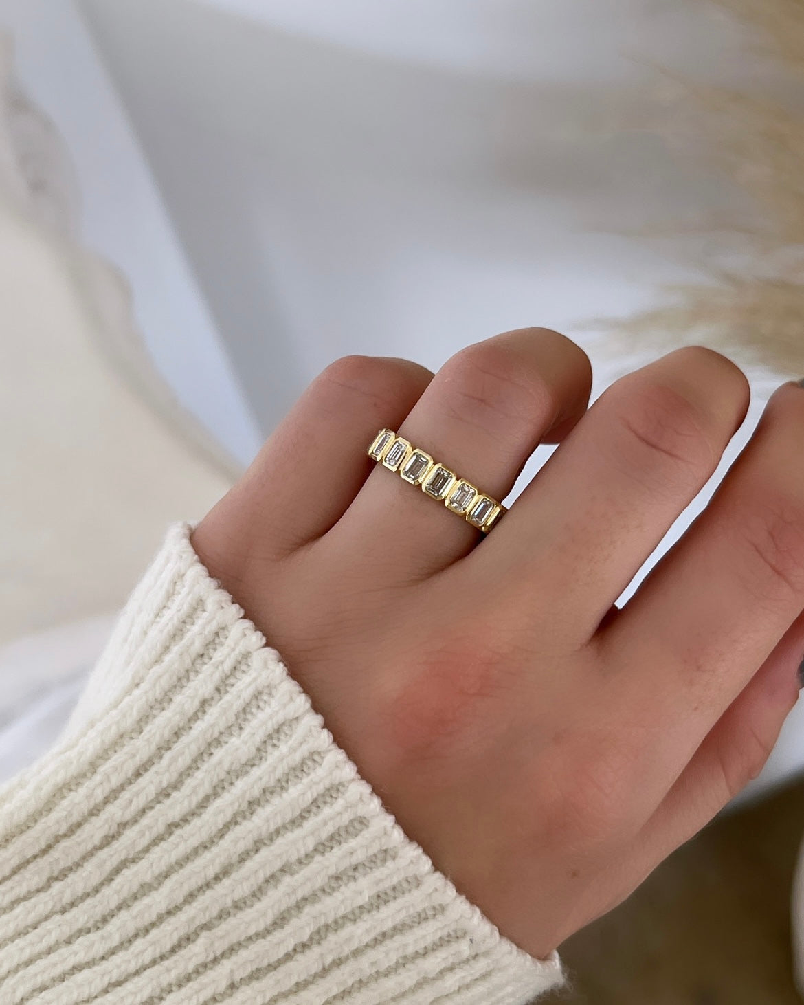 A hand wearing a ribbed white sweater sleeve showcases the Bezel Set Emerald Cut Eternity Ring, made from 18KT gold with rectangular VS clarity gemstones. The softly blurred background highlights the F color brilliance and intricate details of the ring.