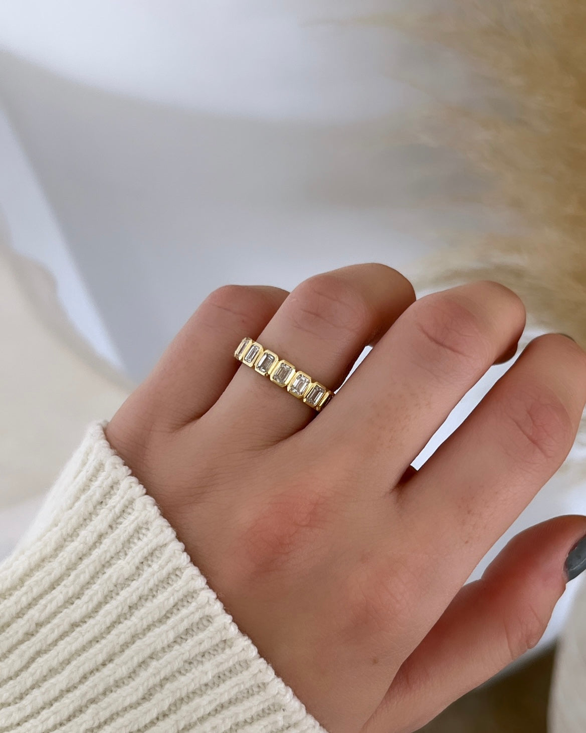 A hand in a ribbed white sweater displays the Bezel Set Emerald Cut Eternity Ring, crafted in 18KT gold with rectangular F Color, VS Clarity gemstones. The softly blurred beige and off-white backdrop enhances the rings elegant sparkle.