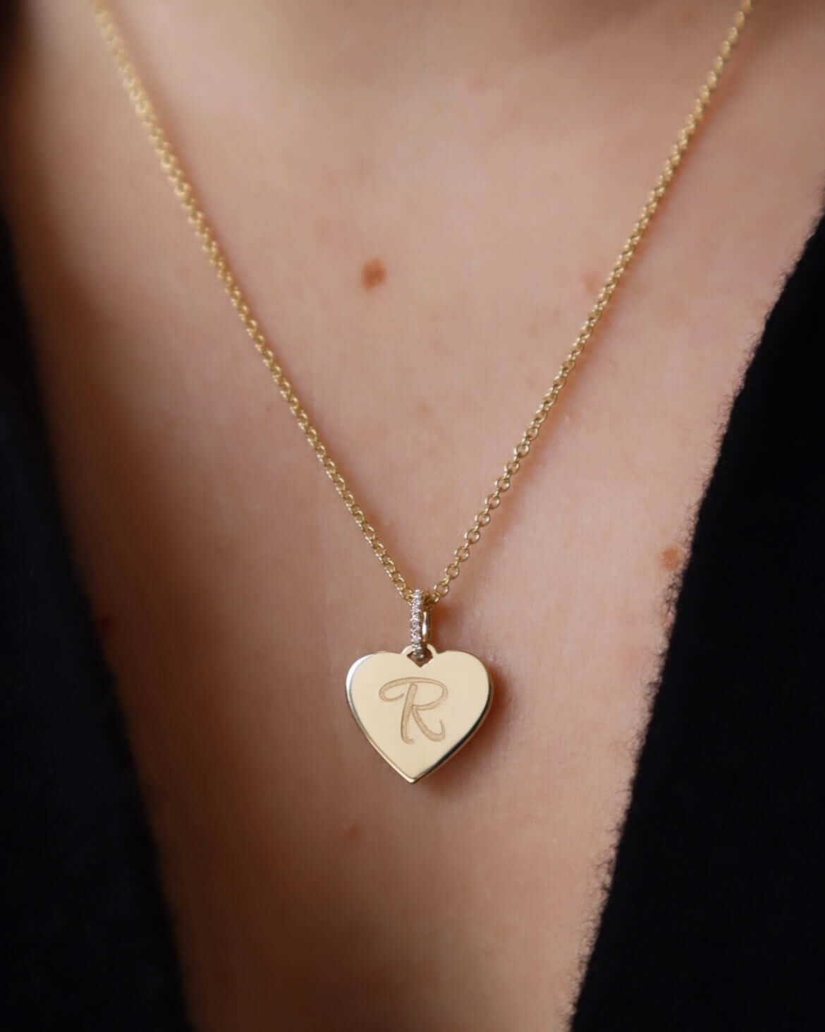 A close-up of the Mini Heart Engravable Pendant Necklace, featuring a gold heart-shaped pendant engraved with the letter R. It hangs from a fine chain around a persons neck, who is wearing a black top.