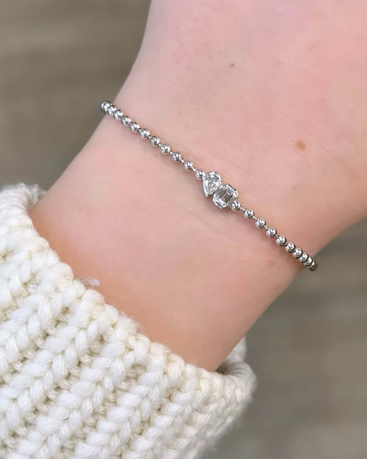 An Emerald & Pear Toi Et Moi Bracelet graces the wrist with its exquisite silver design and sparkling diamond centerpiece, perfectly paired with a cream-colored knitted sweater against a blurred backdrop symbolizing cherished love.