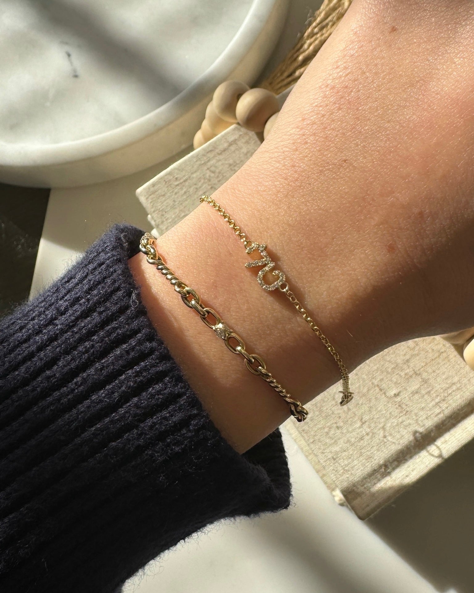 A wrist adorned with two gold bracelets: one with large chain links and the Diamond Block Name Bracelet personalized with LOVE. A dark knitted sleeve, bright sunlight casting soft shadows, and a marble surface partially visible highlight these unique pieces.