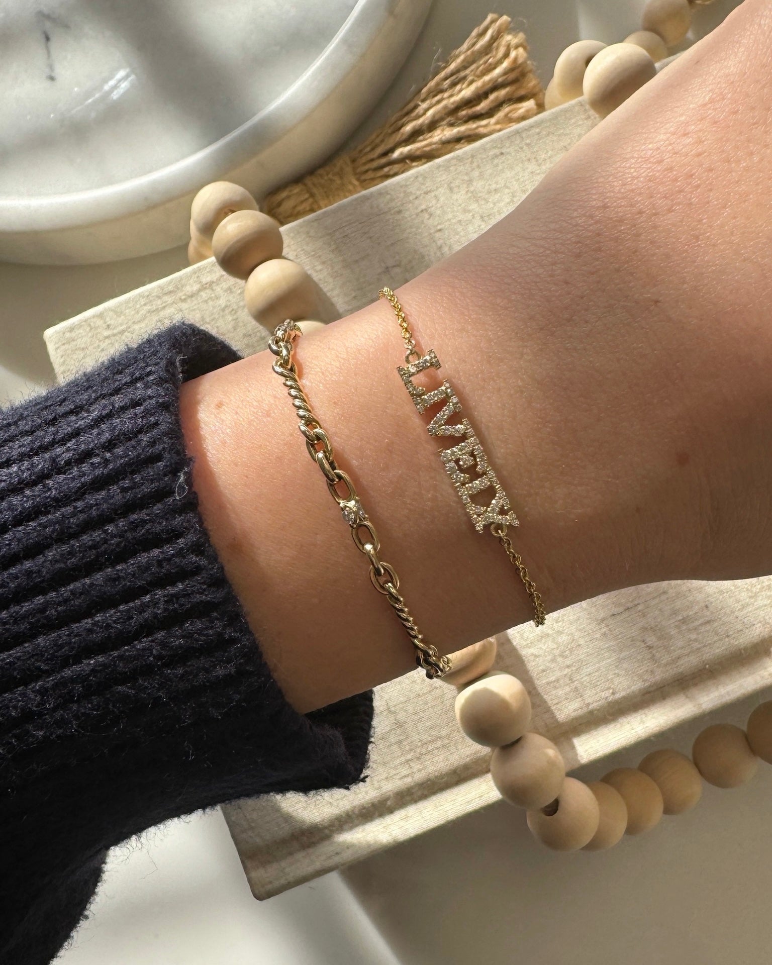 A wrist adorned with two delicate bracelets, featuring a small chain and the Diamond Block Name Bracelet with LIVE in sparkly letters, rests on a beaded item. Soft light casts subtle shadows, creating a truly unique piece of personalized jewelry.