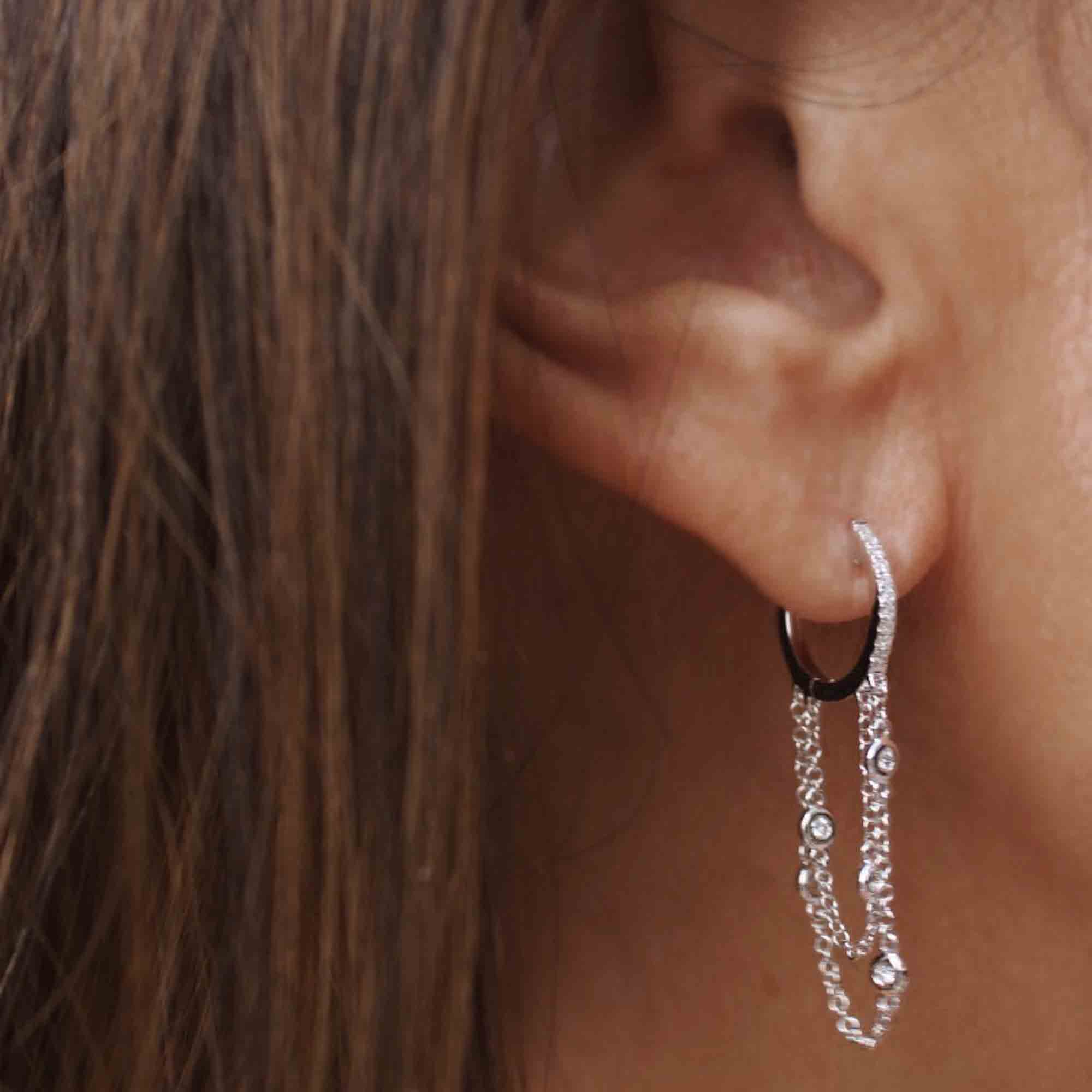 Close-up of a person wearing the Diamond Waterfall Huggies from Janet Heller Fine Jewelry. These small hoop earrings have dangling chains with delicate decorative elements. The persons long brown hair complements the elegant design.