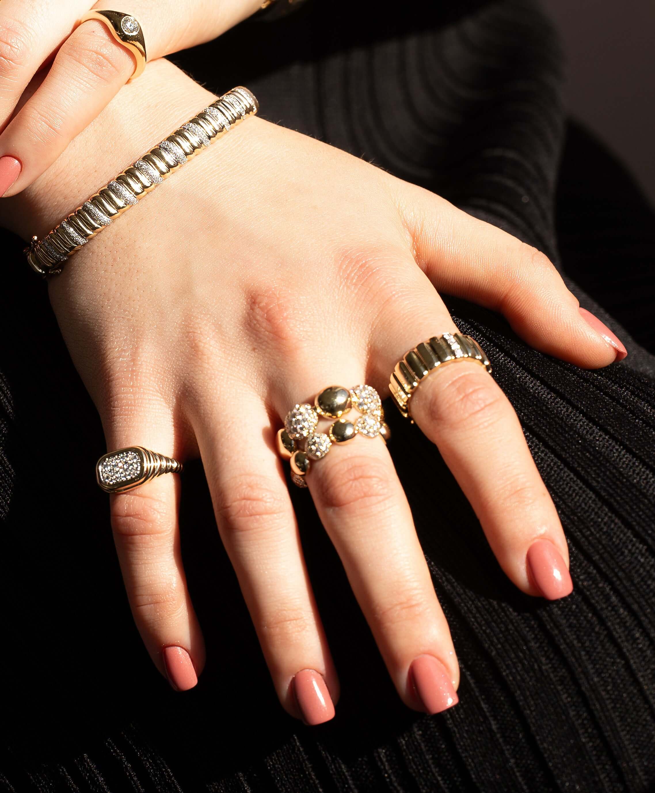A close-up features a hand adorned with multiple gold rings, including the Large Alternating Pave Diamond Bubble Ring, showcasing fine jewelry designs. A bracelet graces the wrist, with nails in soft pink. The hand elegantly rests on textured black fabric, inviting preorders for these exquisite pieces.
