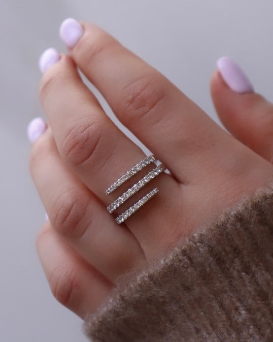 A hand with light purple nail polish displays our Diamond Spiral Ring, featuring three parallel diamond-studded bands on the ring finger. A cozy brown fuzzy sweater sleeve enhances this elegant preorder essential.