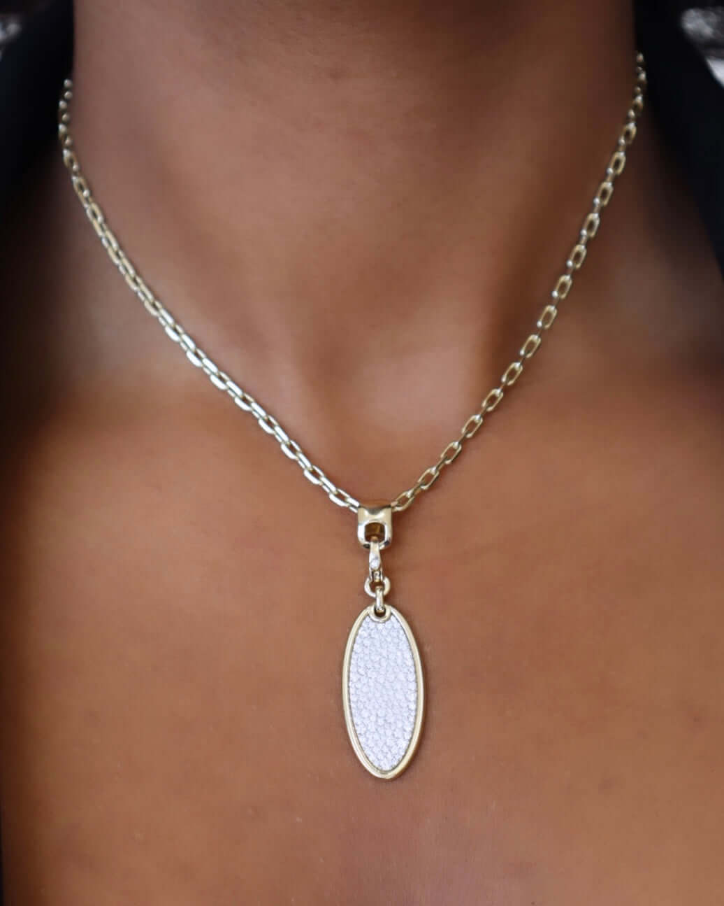A close-up shows a person wearing the Oval Diamond Charm from Janet Heller Fine Jewelry, featuring a silver chain and an oval pendant encrusted with small white gems. The pendant sits centrally on the chest against a blurred background of skin and collar.