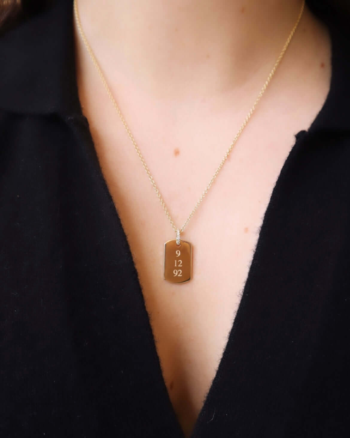 Worn by a person in a black V-neck top, the Large Dog Tag Engravable Pendant Necklace—a striking gold piece featuring a rectangular pendant engraved with the numbers 9 12 92—makes for stunning personalized jewelry.