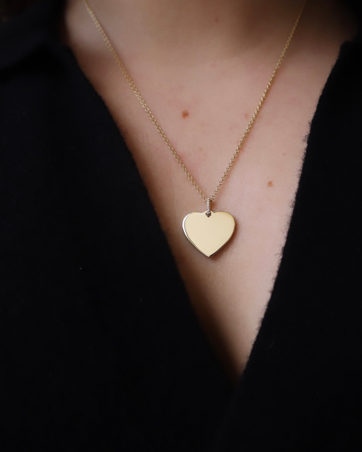The Large Heart Engraveable Pendant Necklace, a stunning gold heart-shaped piece with personalized engraving on a delicate chain, graces the neck. Its refined elegance and shiny design contrast beautifully with the black fabric backdrop, making this engraved keepsake truly captivating.