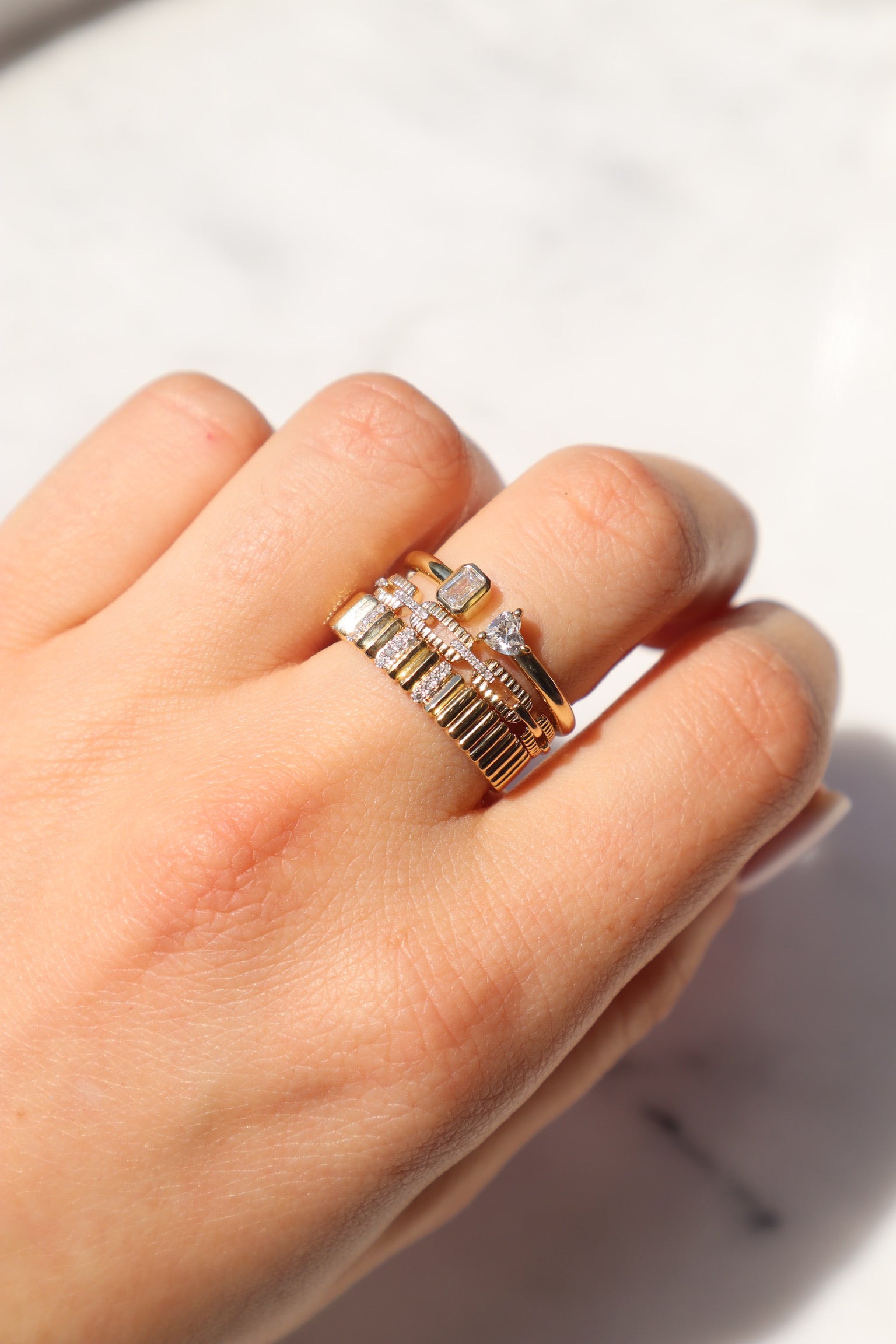 On a pristine white background, a hand showcases two elegant Emerald & Heart Toi Et Moi rings adorned with small rectangular and square diamond-like stones, embodying a timeless love story.