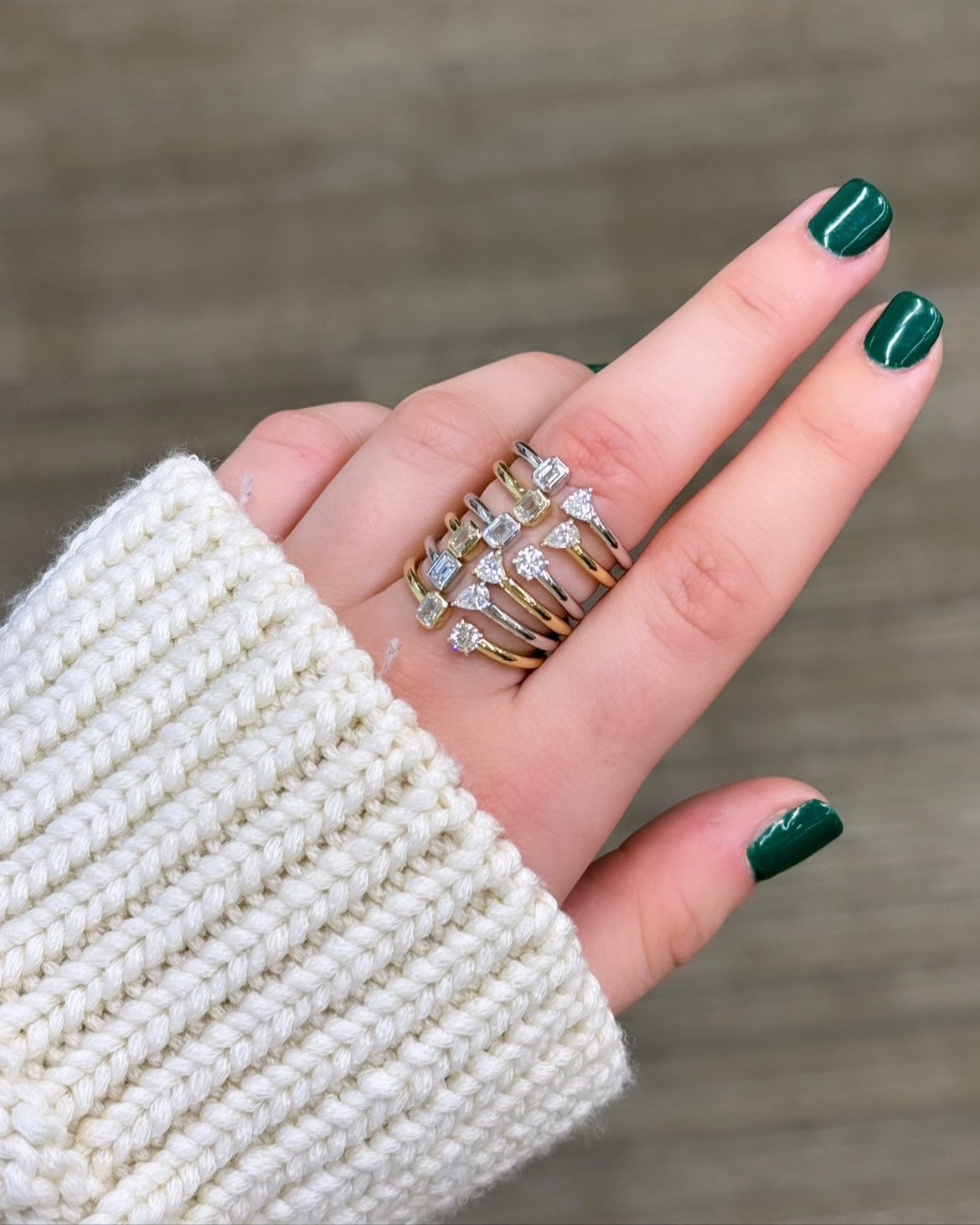 A hand with green painted nails, dressed in a knitted white sweater, wears multiple gold rings including an Emerald & Pear Toi Et Moi Ring that symbolizes love, adorned with small diamonds on two fingers.