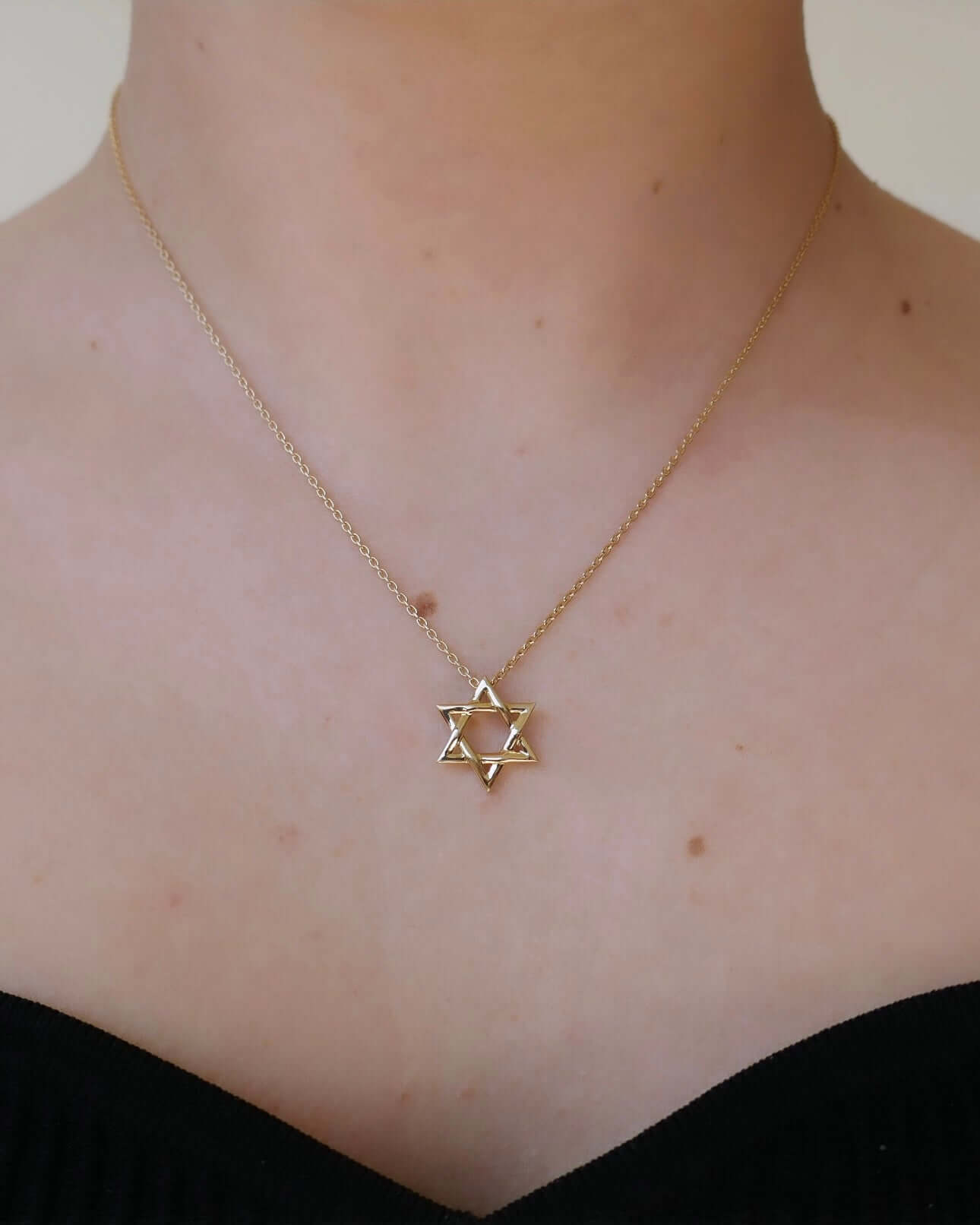 Wearing the Star of David Necklace, featuring a delicate gold chain and subtle diamond accents, a person exudes elegance as it rests gracefully on their skin above a black garment against a neutral background.