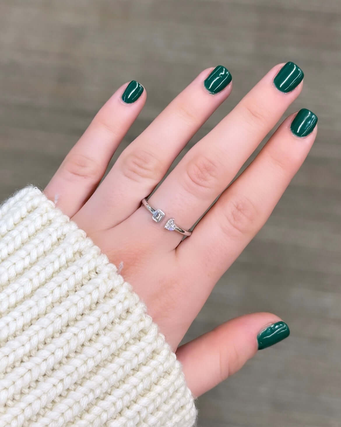 A hand with short, dark green polished nails wears the Emerald & Heart Toi Et Moi Ring in silver, adorned with two stones. A cream-colored knitted sweater sleeve partially covers the wrist, suggesting cozy love stories against a soft, neutral background.