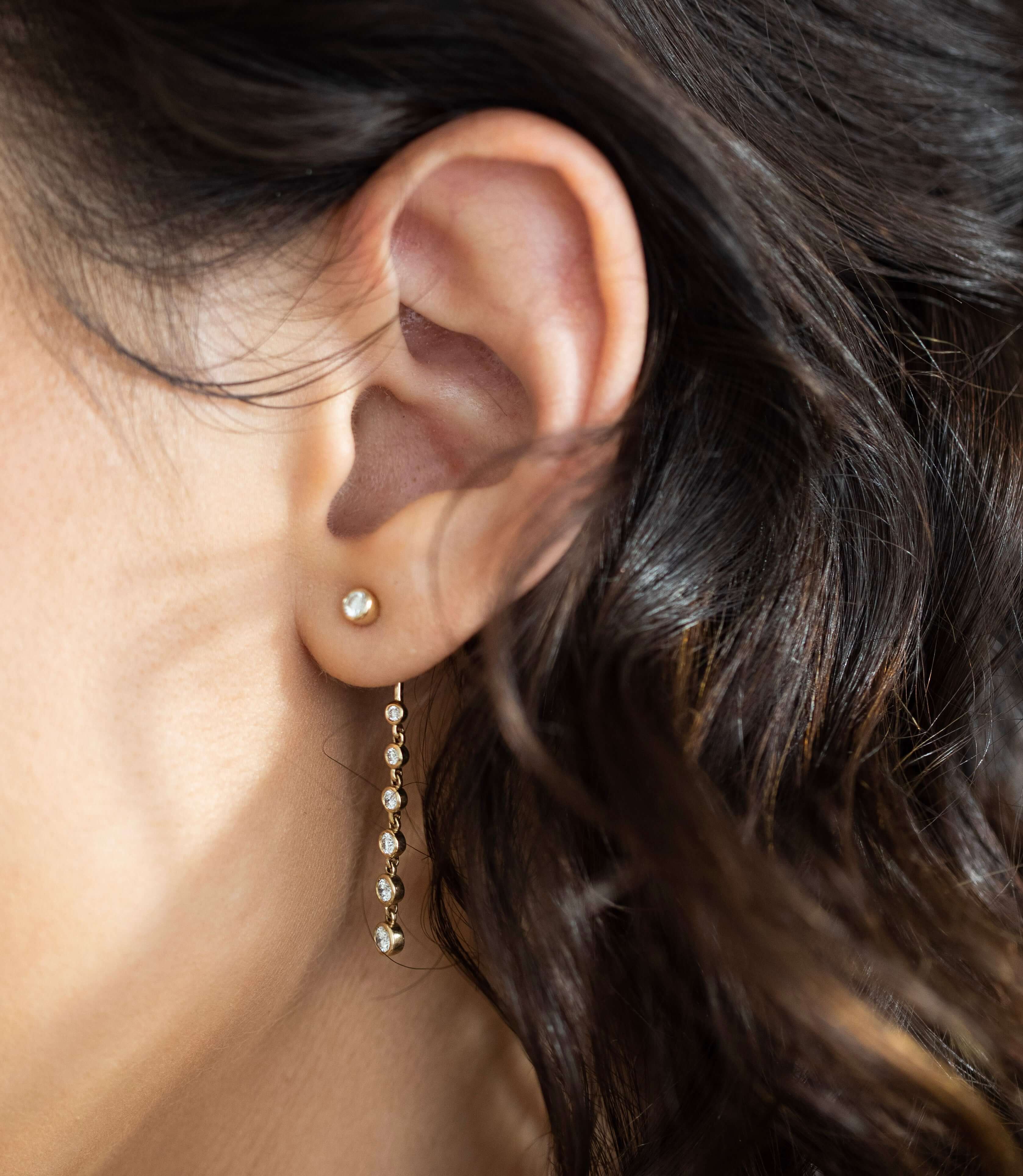 Close-up of a person's ear adorned with two earrings: a small gold stud and the Diamond Jacket Drop Earrings from Janet Heller Fine Jewelry, featuring an elegant gold drop design with multiple shimmering beads. The person’s dark, wavy hair partially frames the ear, highlighting the sophistication of this custom jewelry piece. For inquiries about the Diamond Jacket Drop Earrings or other fine jewelry, please contact Janet Heller Fine Jewelry.