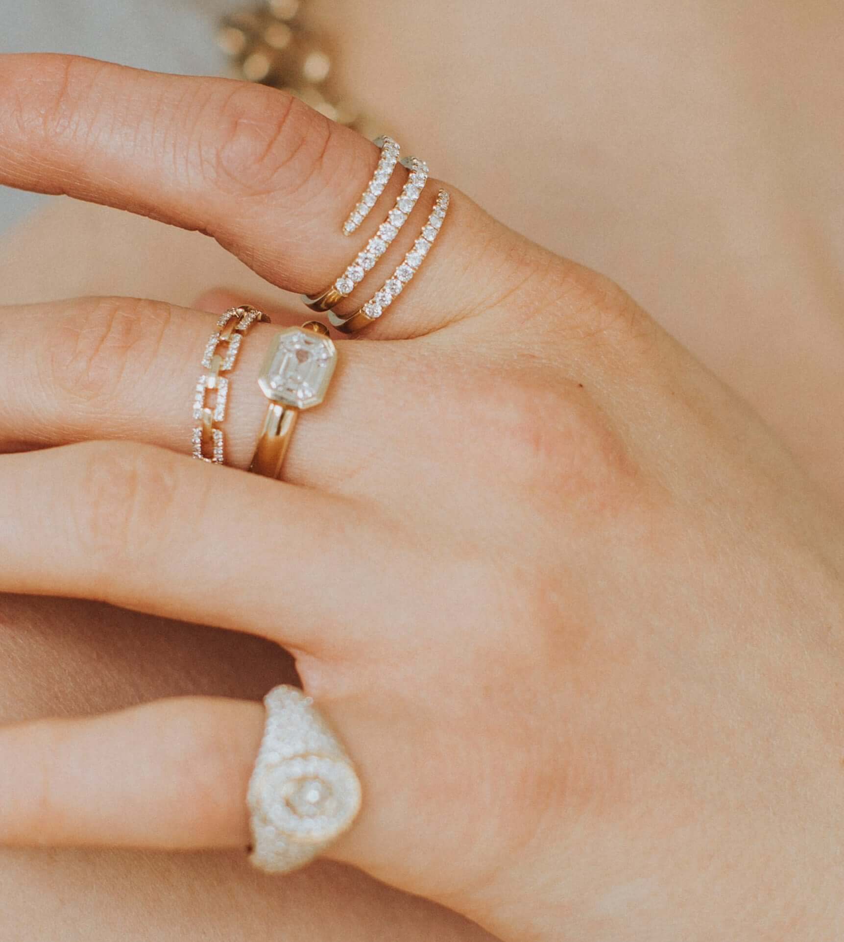 A close-up highlights a hand wearing the Diamond Athena Ring and an emerald-cut gem. Set against fair skin and a softly blurred background, the rings glisten, inviting admiration and possibly inspiring a preorder for lovers of elegance.