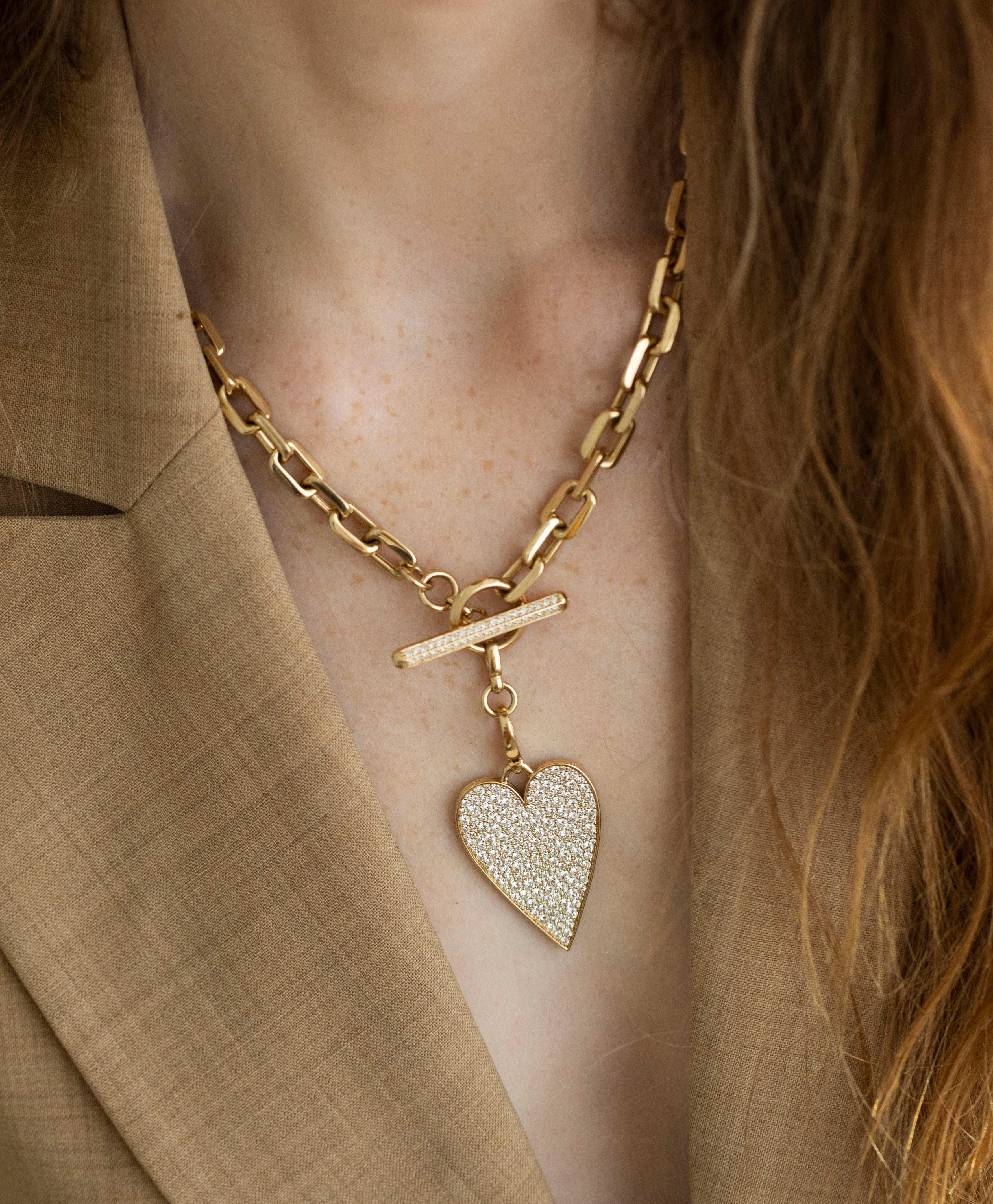 Close-up of a person wearing Janet Heller Fine Jewelrys gold chain necklace with the Jumbo Flat Diamond Heart Charm over a brown blazer. The toggle clasp enhances its customization, while their long hair cascades to the right.