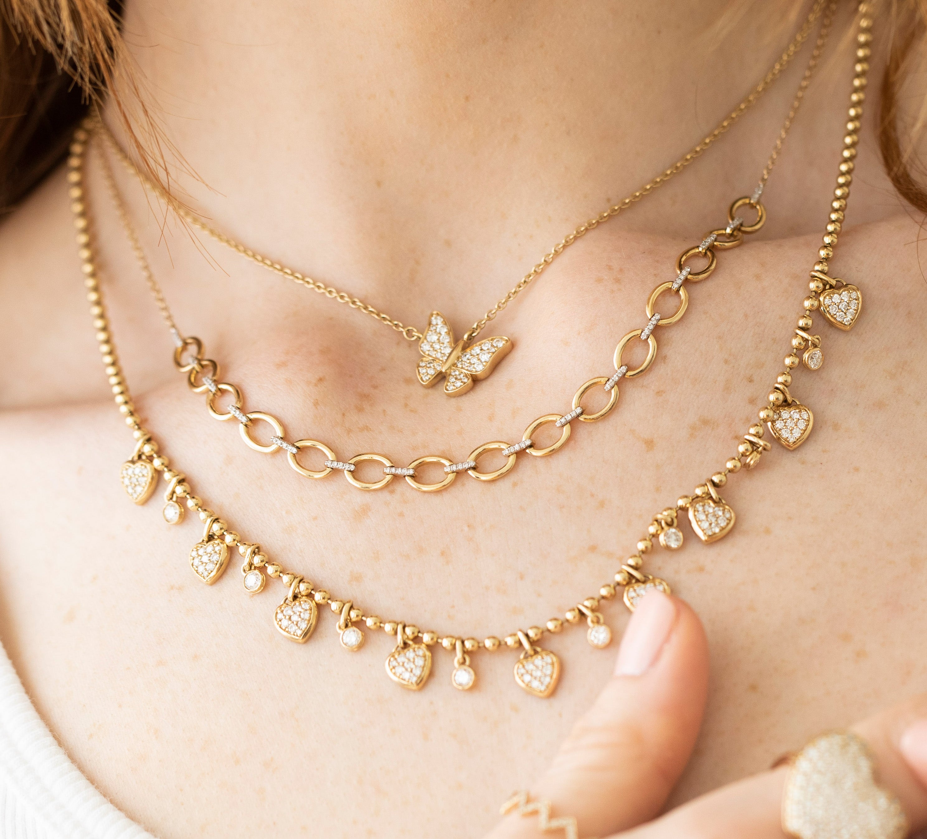 Close-up of a person wearing three exquisite gold necklaces: the top is a Mini Sideways Butterfly Necklace, the middle has a chain-link design, and the lowest features heart-shaped pendants. This stunning ensemble enhances any look, perfectly complementing a white top.