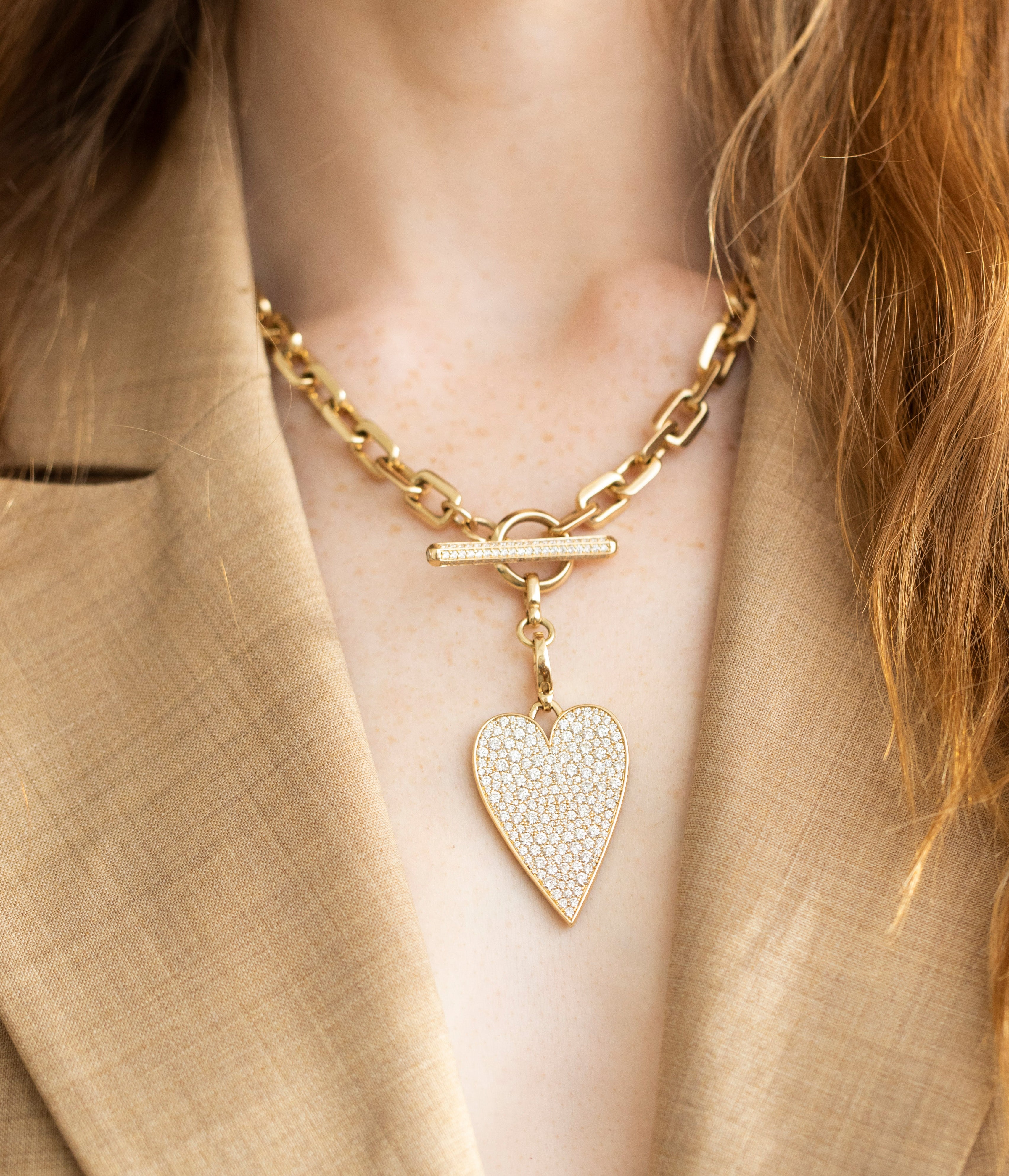 A woman in a beige blazer showcases the Janet Heller Fine Jewelry Chunky Diamond Toggle Link Necklace, featuring a large heart-shaped pendant with small crystal charms.