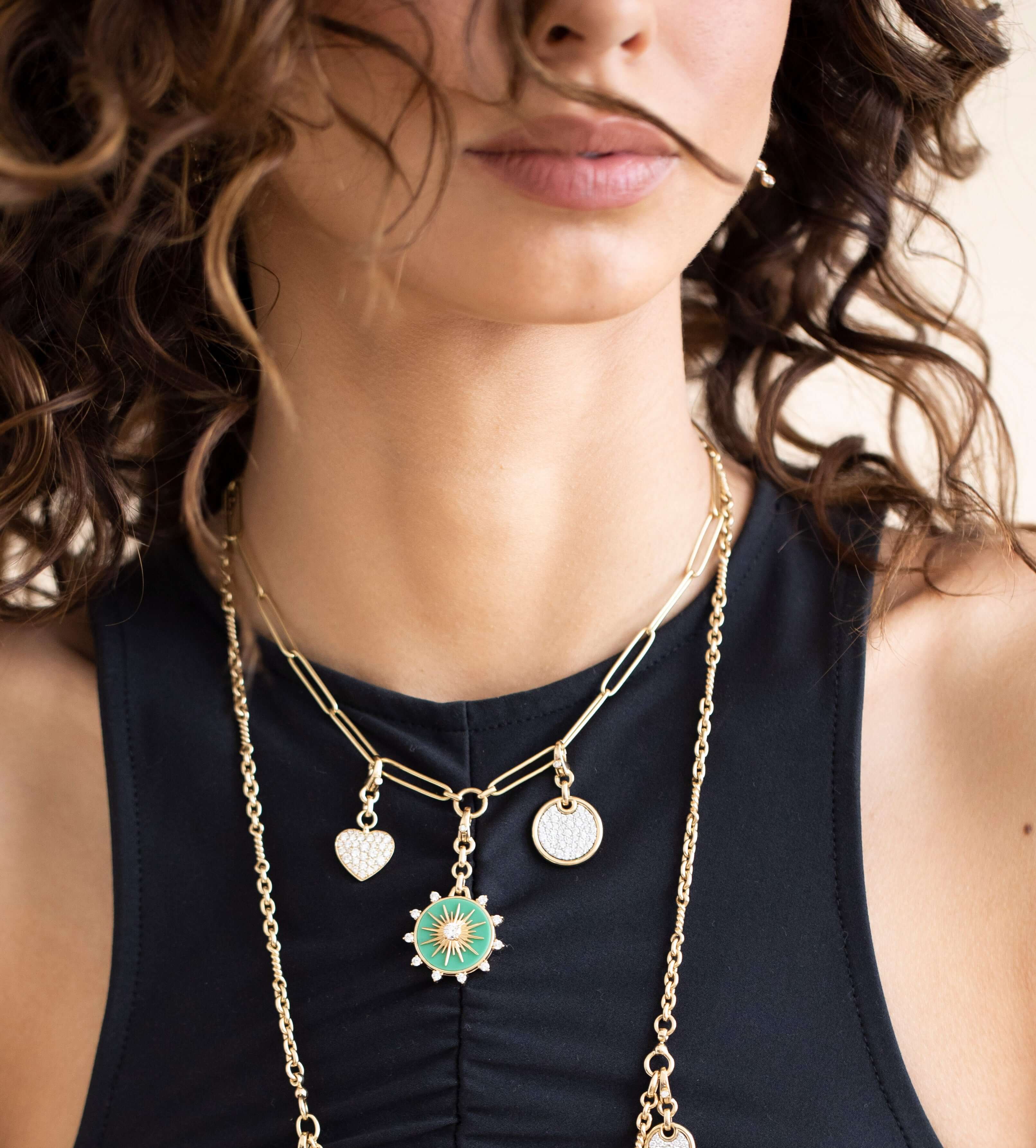 A woman with curly hair wears a black sleeveless top and layered gold necklaces from Janet Heller Fine Jewelry. One necklace features a chrysoprase starburst pendant, while another has custom charms, including a heart-shaped charm. Her face is partially obscured by her hair.