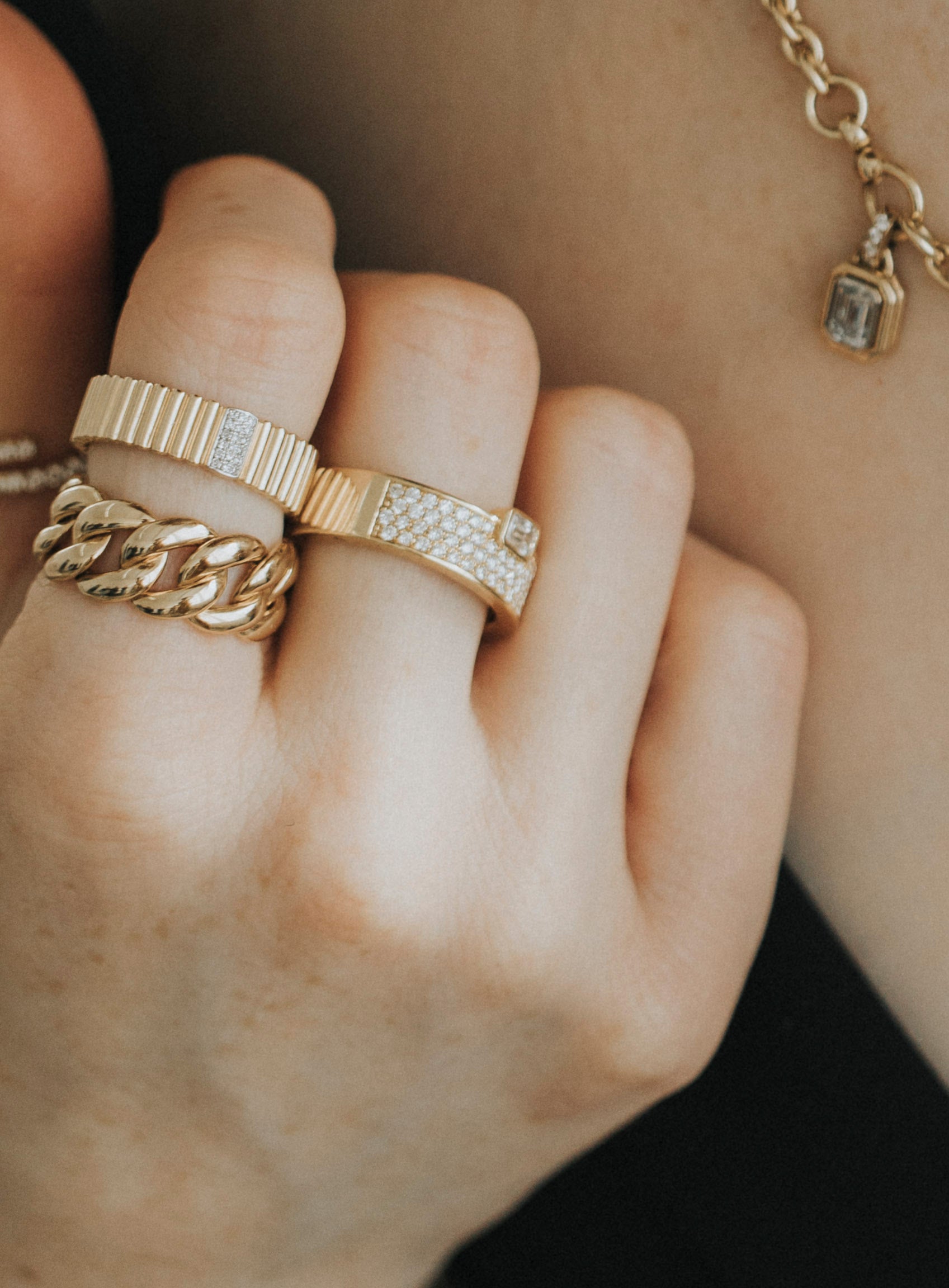 A hand is adorned with three stunning gold rings, featuring the Diamond Sloane Ring, a chain-like design, and a textured pattern. A delicate gold bracelet with a rectangular charm graces the wrist for preorder against an elegantly blurred background.
