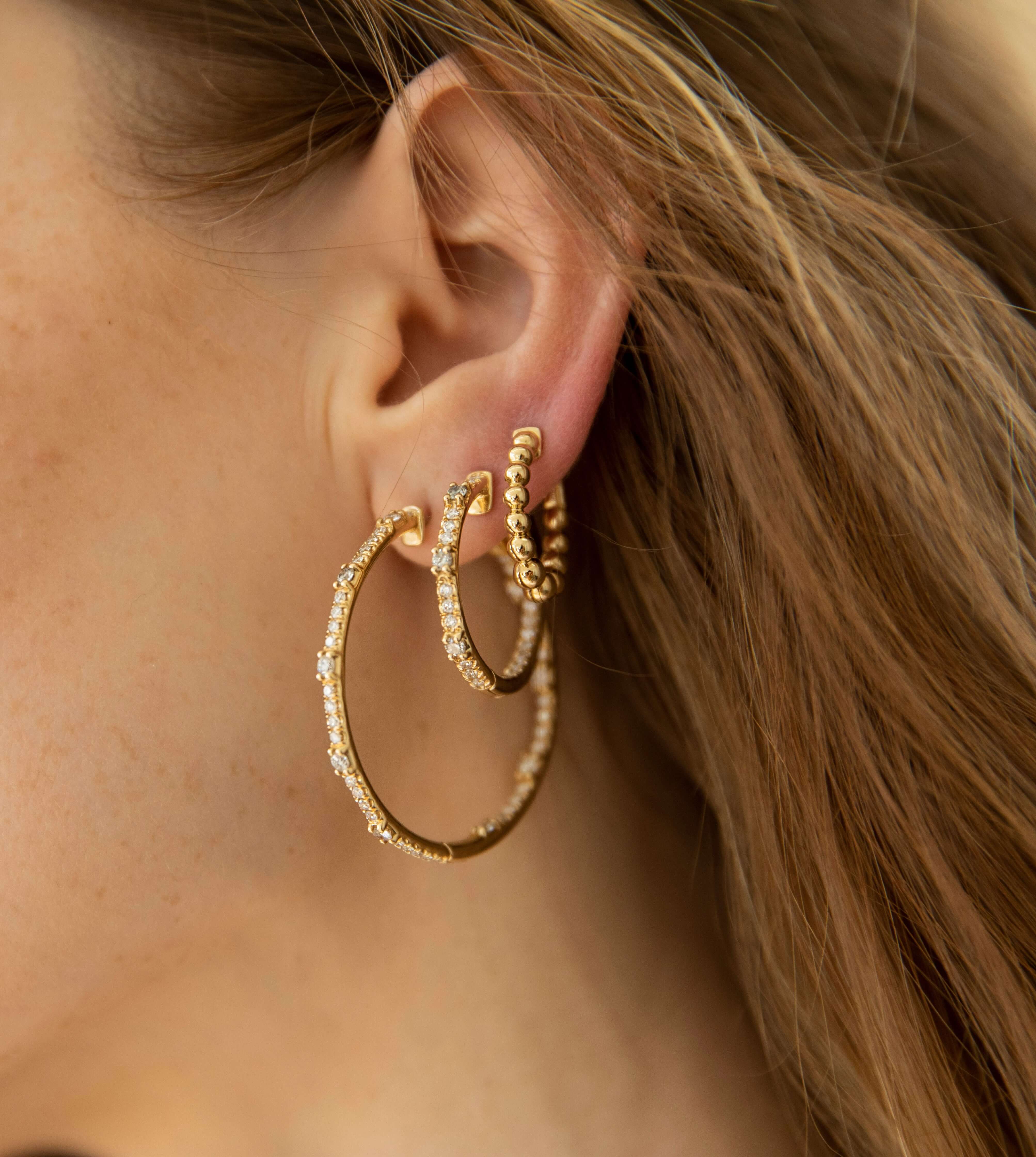 Close-up of a woman's ear adorned with three Mini Nora Hoops from Janet Heller Fine Jewelry (SKU: EAR-1173-Y), each crafted in gold and set with small clear gemstones, against a softly blurred background of light brown hair. For custom jewelry or inquiries, please contact us.